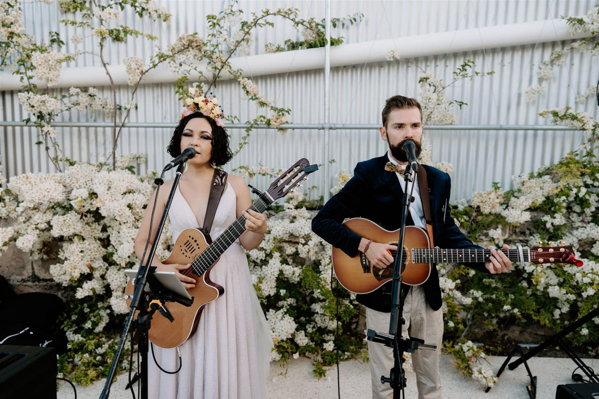 Acoustic duo performing live with guitars and microphones in front of flowering greenery at a wedding.