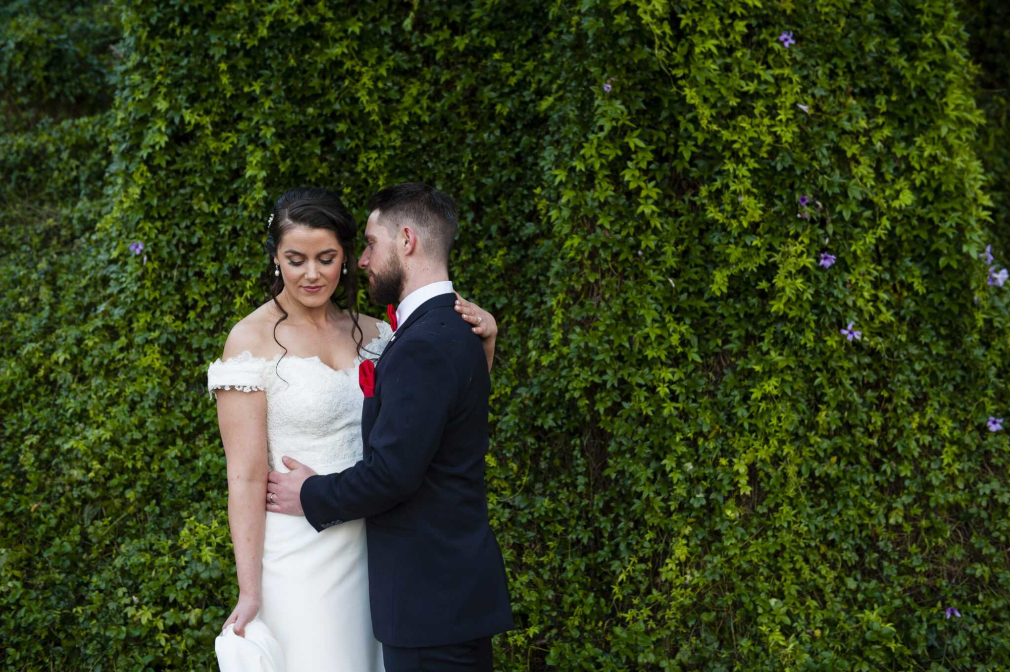 Bride and groom embrace in front of a lush green ivy wall during their outdoor wedding.