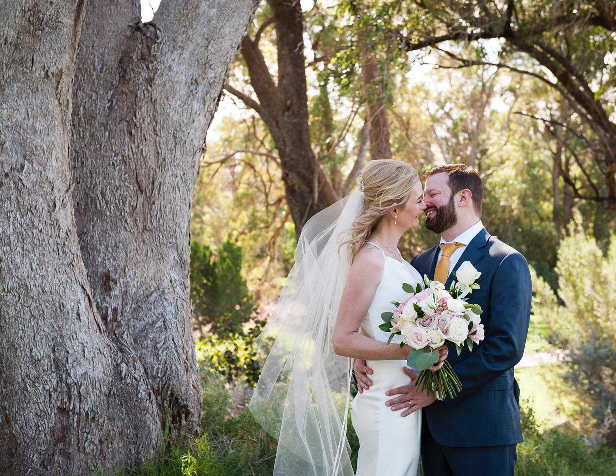 Bride and groom embrace with a pastel bouquet under tall trees in a sunlit outdoor setting.