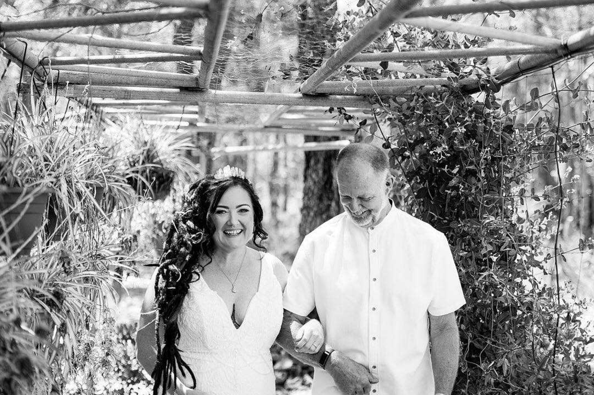 Bride walks arm in arm with her father under a rustic vine-covered garden arbor.