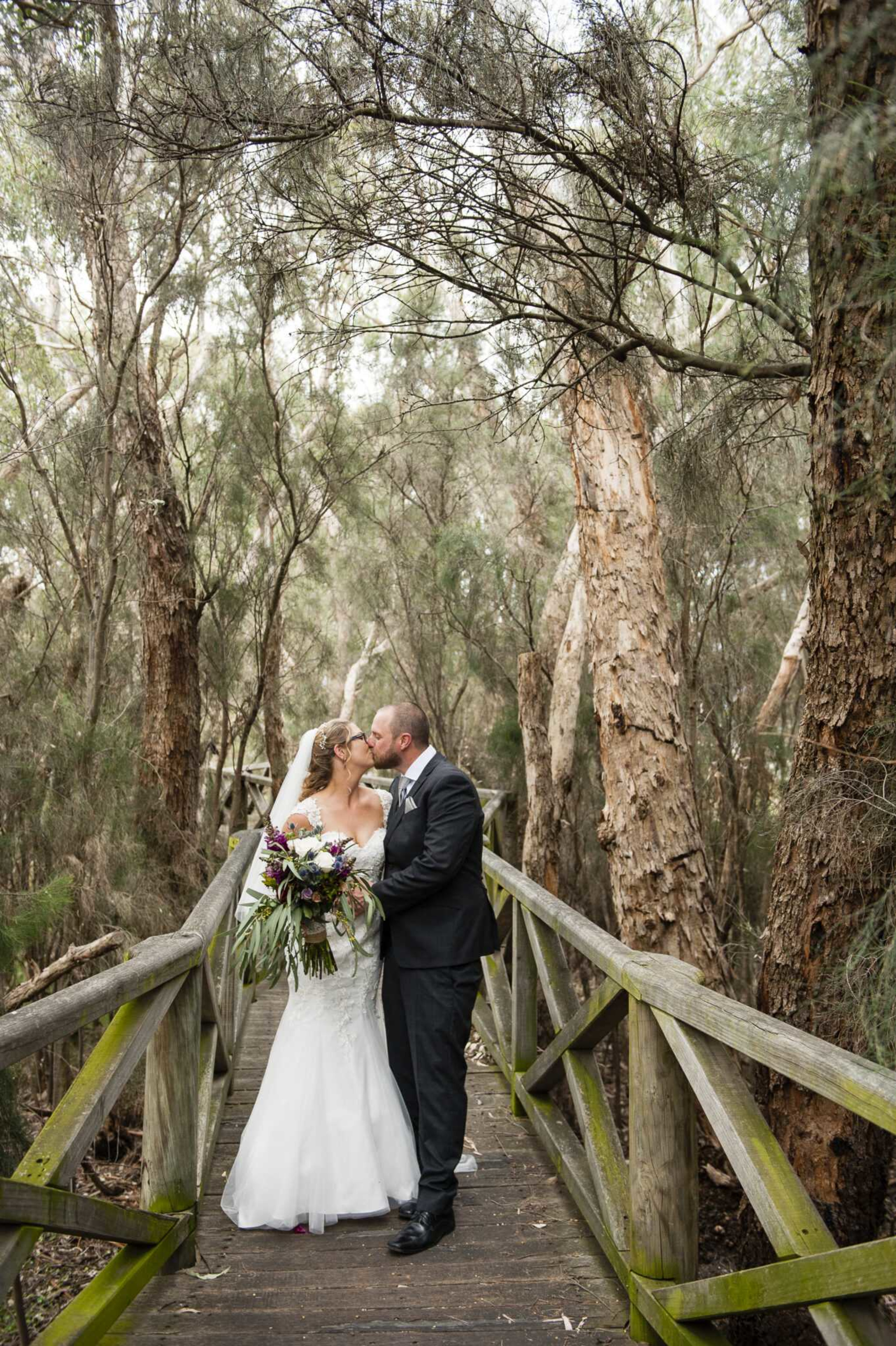 Bride and groom share a kiss on a wooden bridge surrounded by tall forest trees.