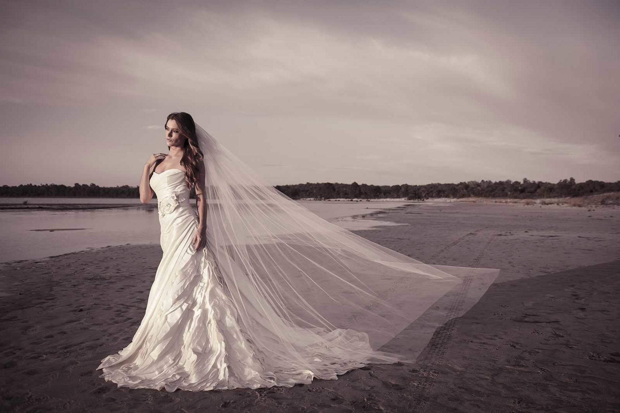 Bride in a flowing strapless gown with a long veil standing on a quiet beach at sunset.