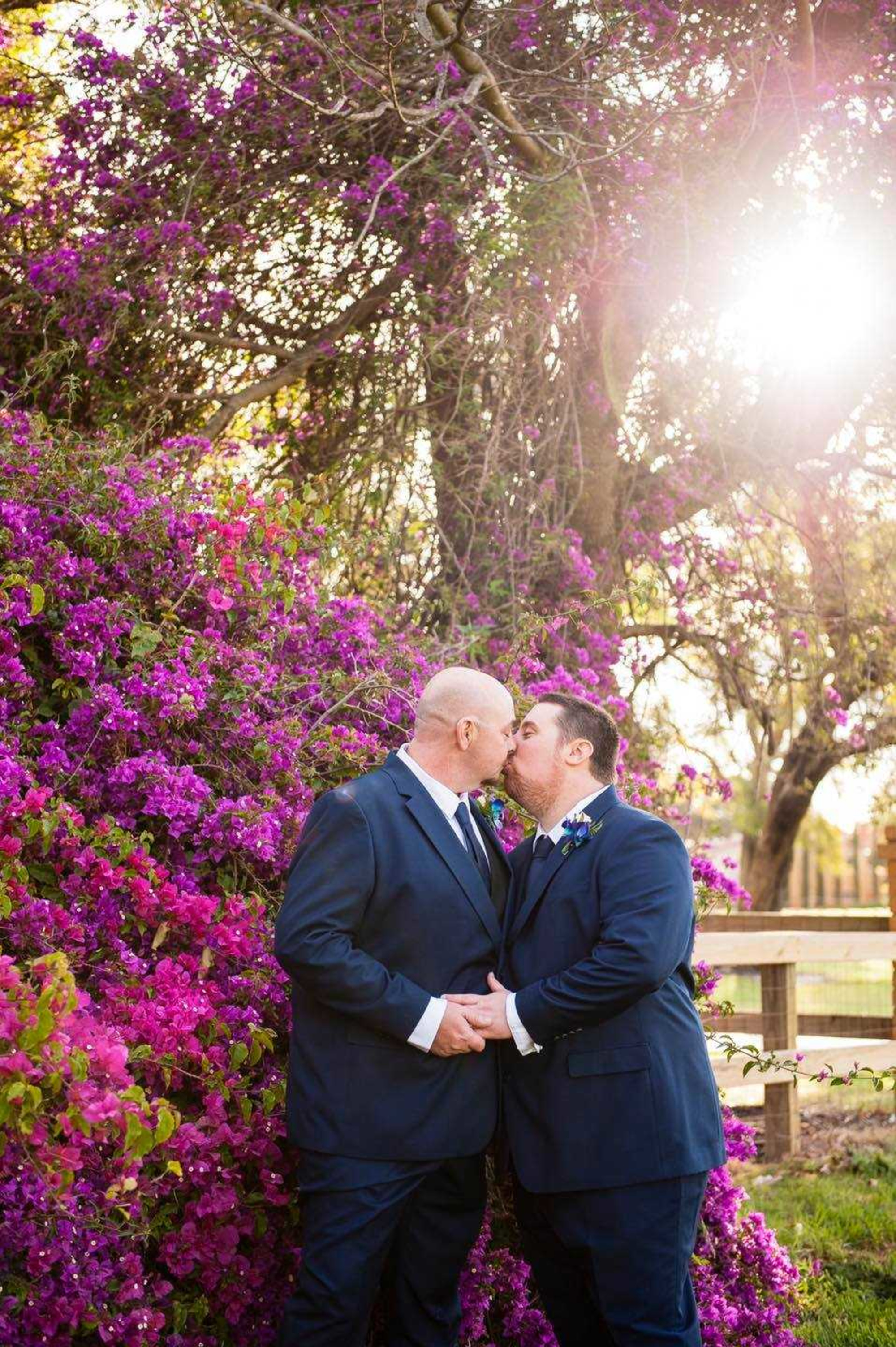 Two grooms in navy suits share a kiss in front of vibrant purple flowers in a sunlit garden.