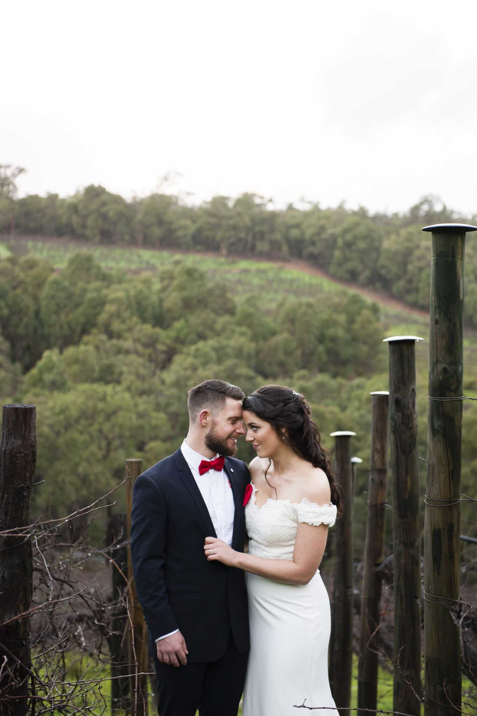 Bride and groom embrace in a vineyard surrounded by green hills and wooden posts.