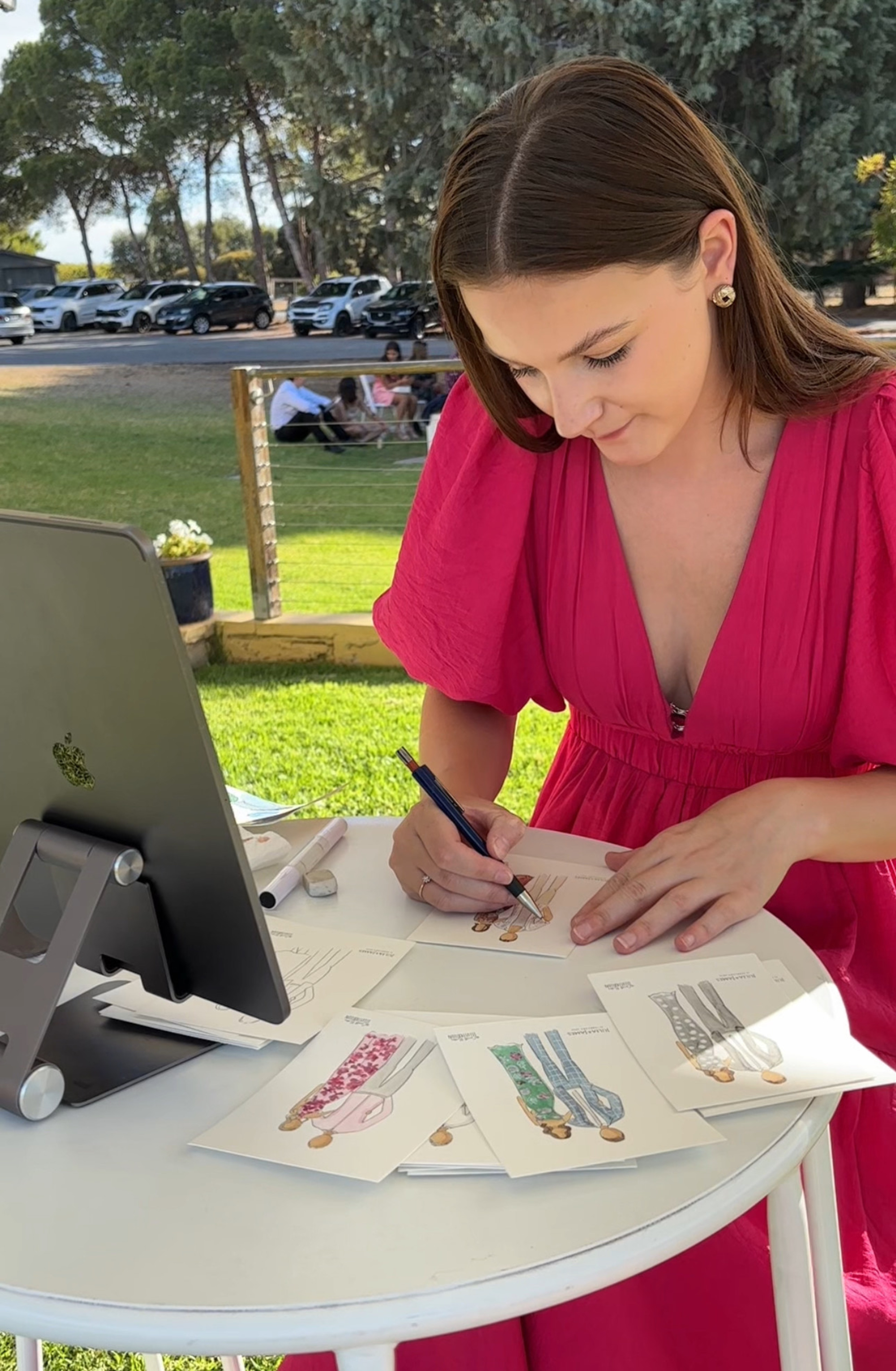 Live wedding artist in a pink dress sketching fashion-style guest portraits at an outdoor reception table.