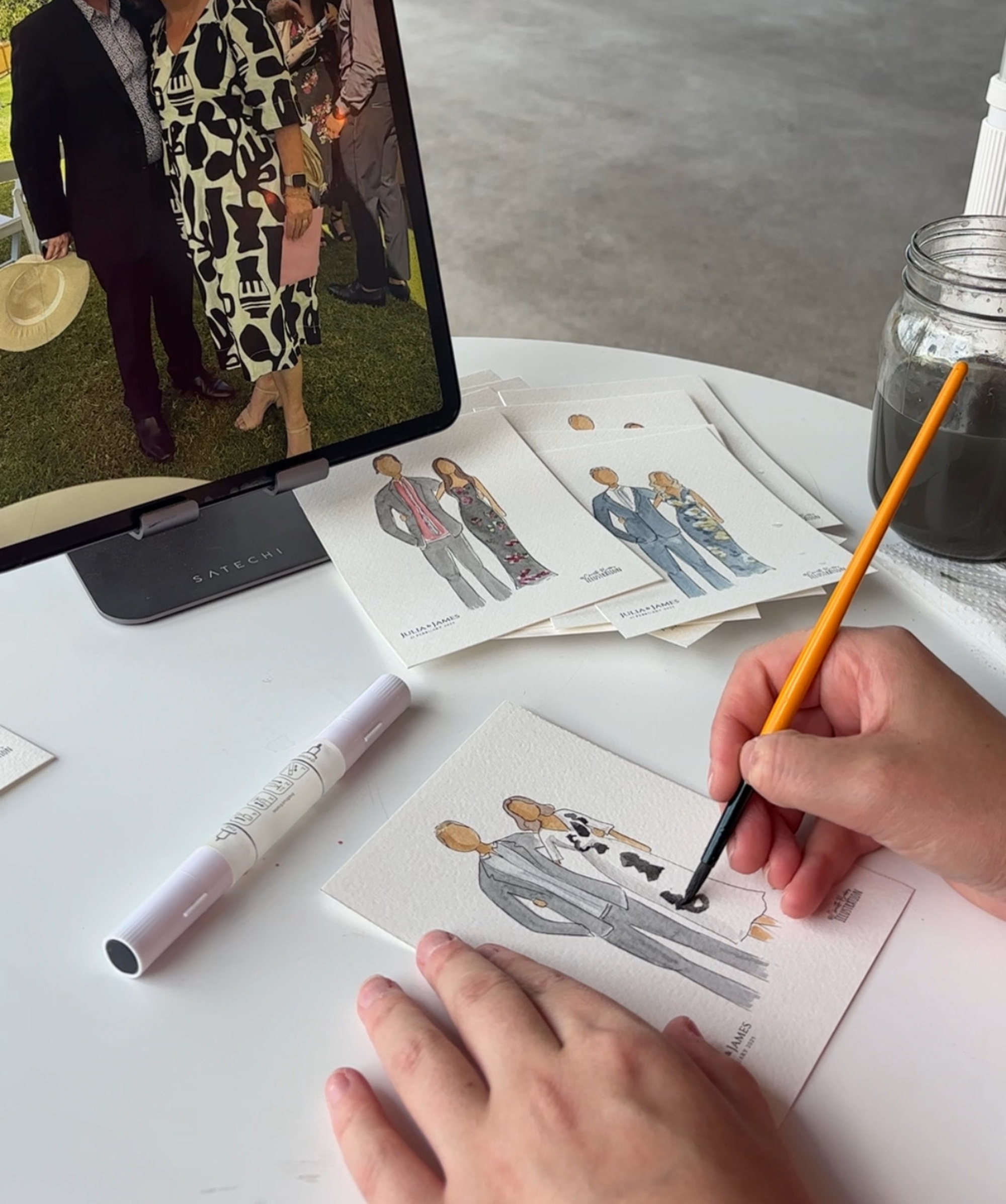 Artist paints custom watercolor portraits of wedding guests from a tablet photo at a white table.