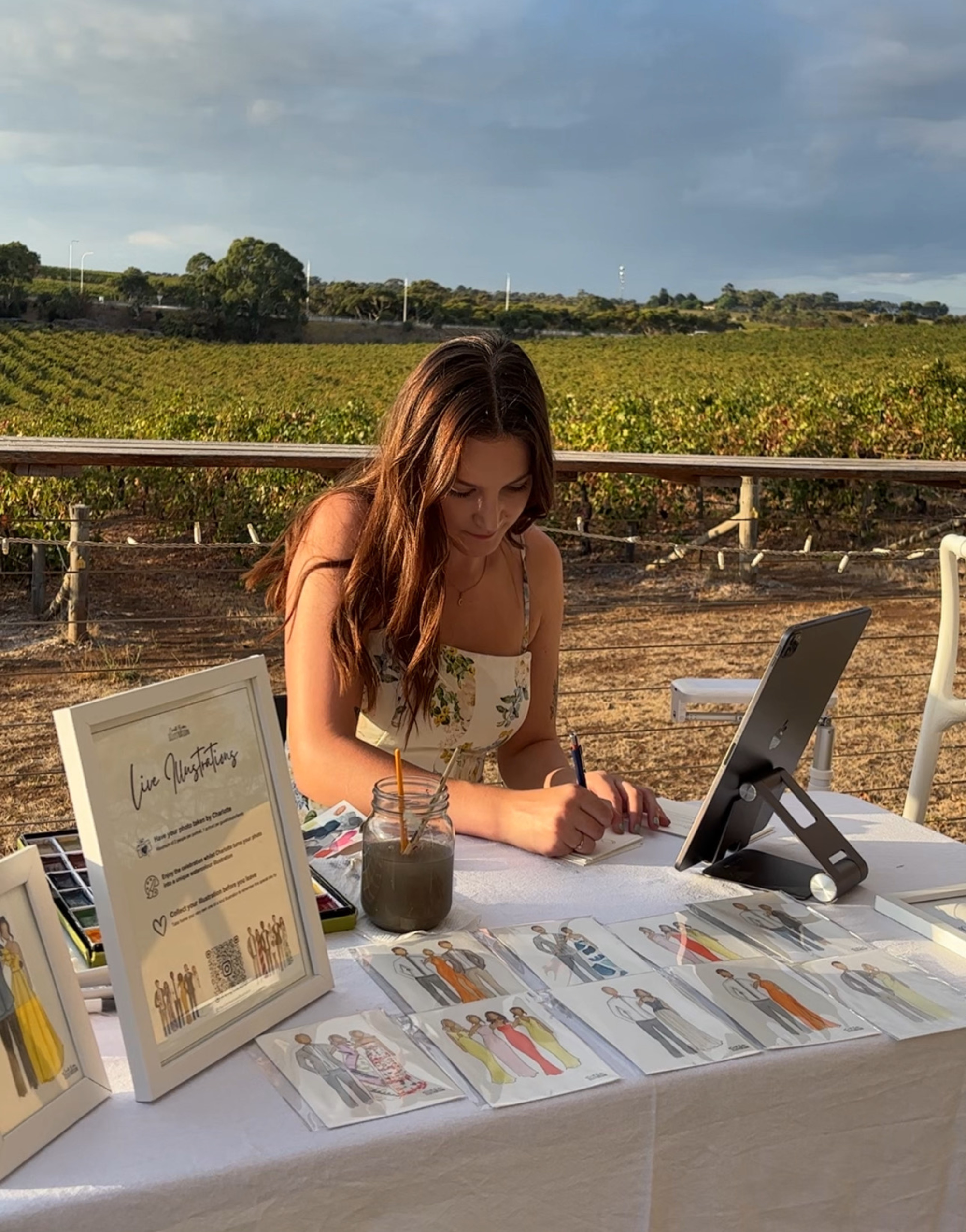 Live illustrator creates custom guest portraits at an outdoor vineyard wedding reception table.