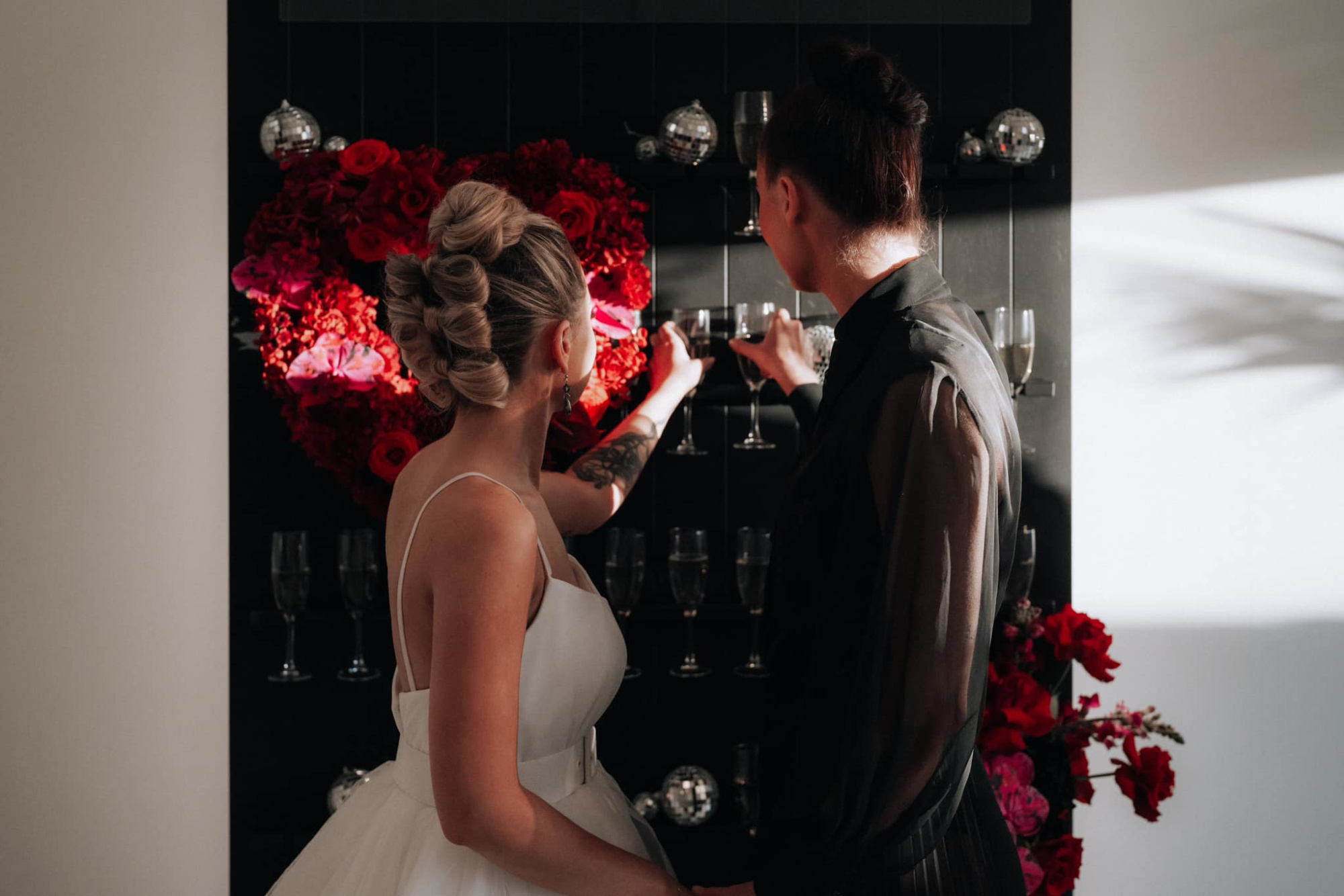 Two brides take champagne glasses from a modern wall display with a red rose heart backdrop.