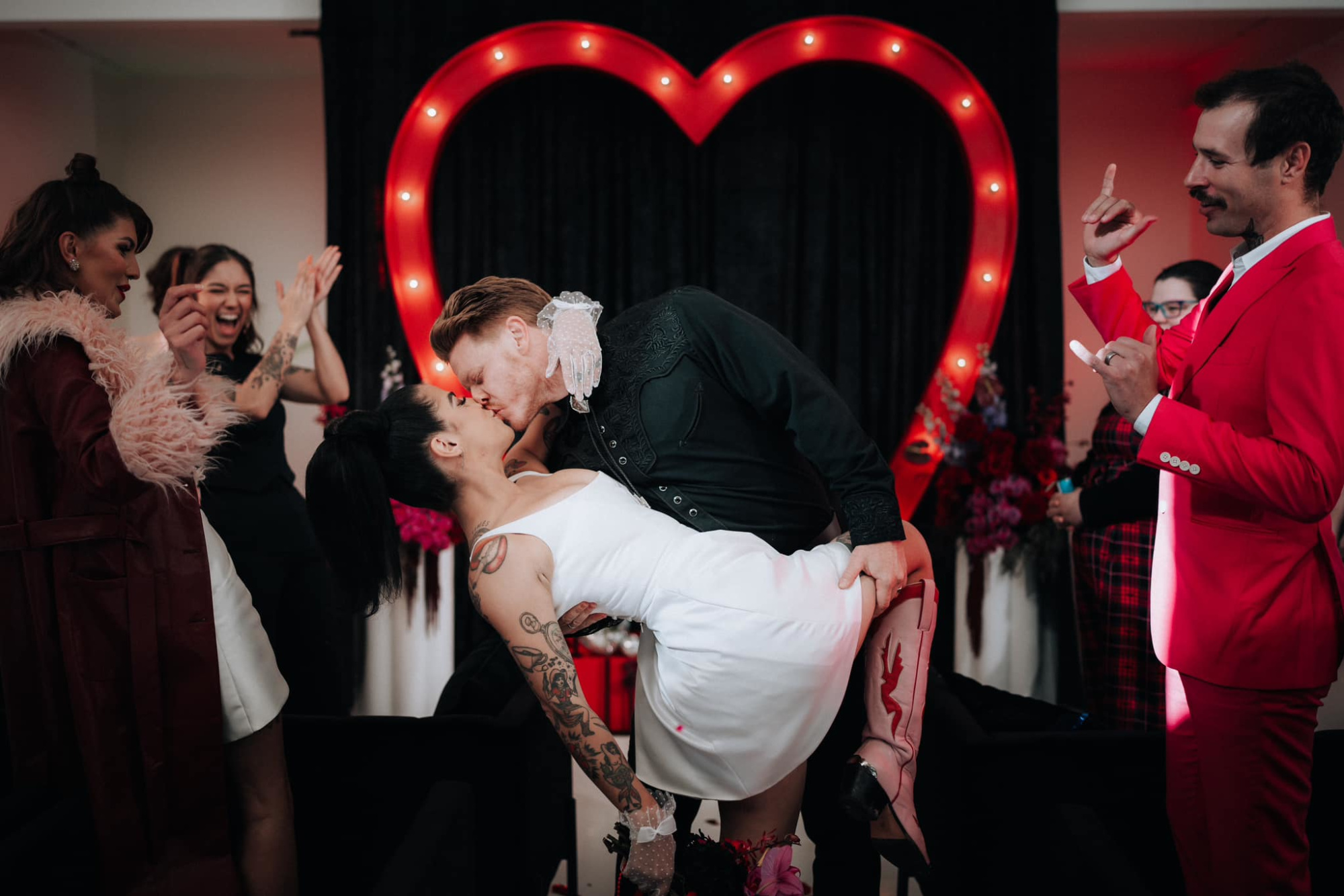 Tattooed couple shares a dramatic kiss under a glowing red heart backdrop as guests cheer around them.