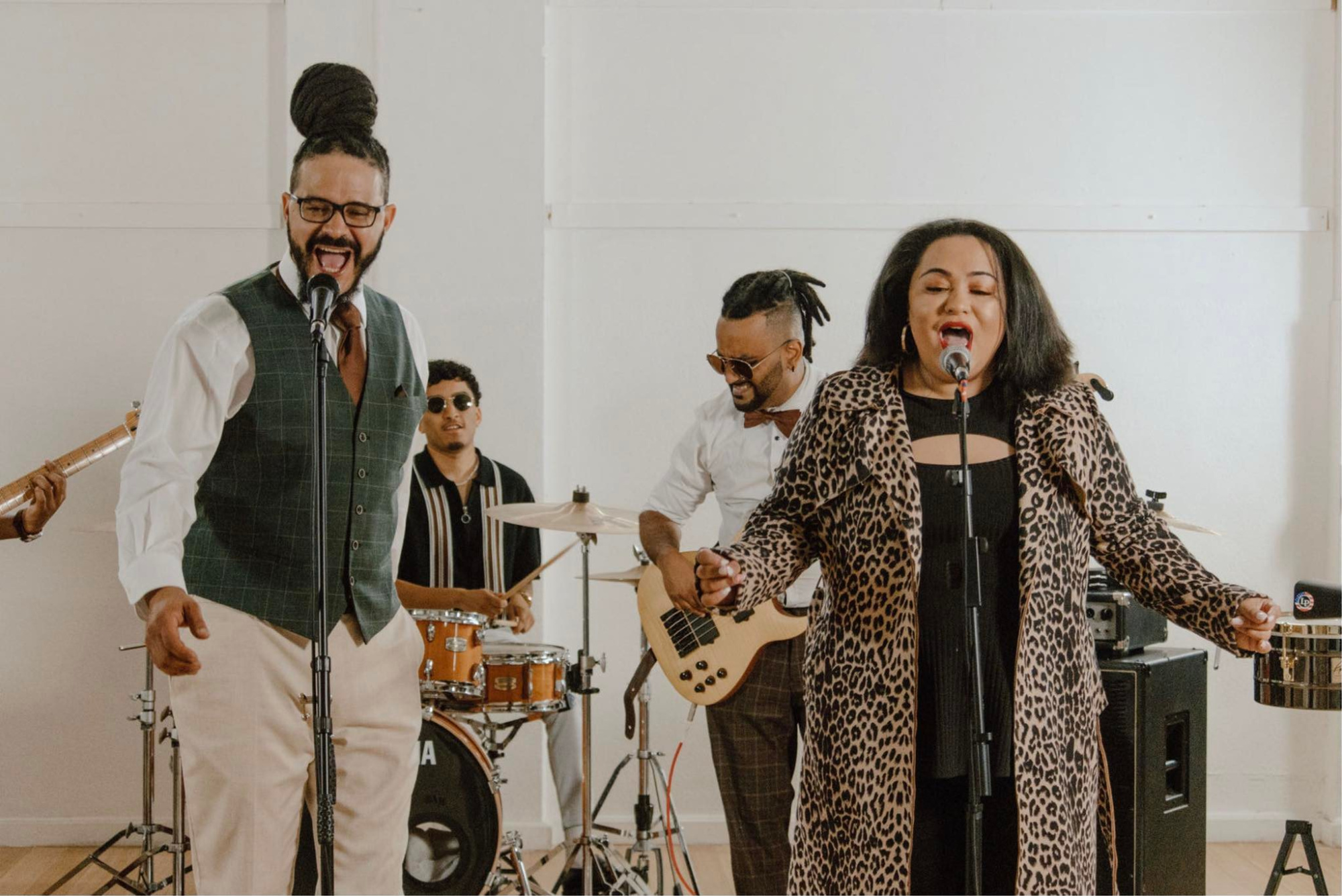 A lively wedding band performs indoors with two vocalists singing into microphones and musicians playing drums and guitar.