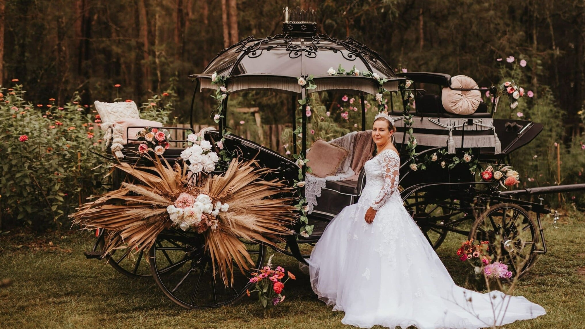 Bride in lace gown posing beside a flower-adorned vintage carriage in a rustic garden setting.