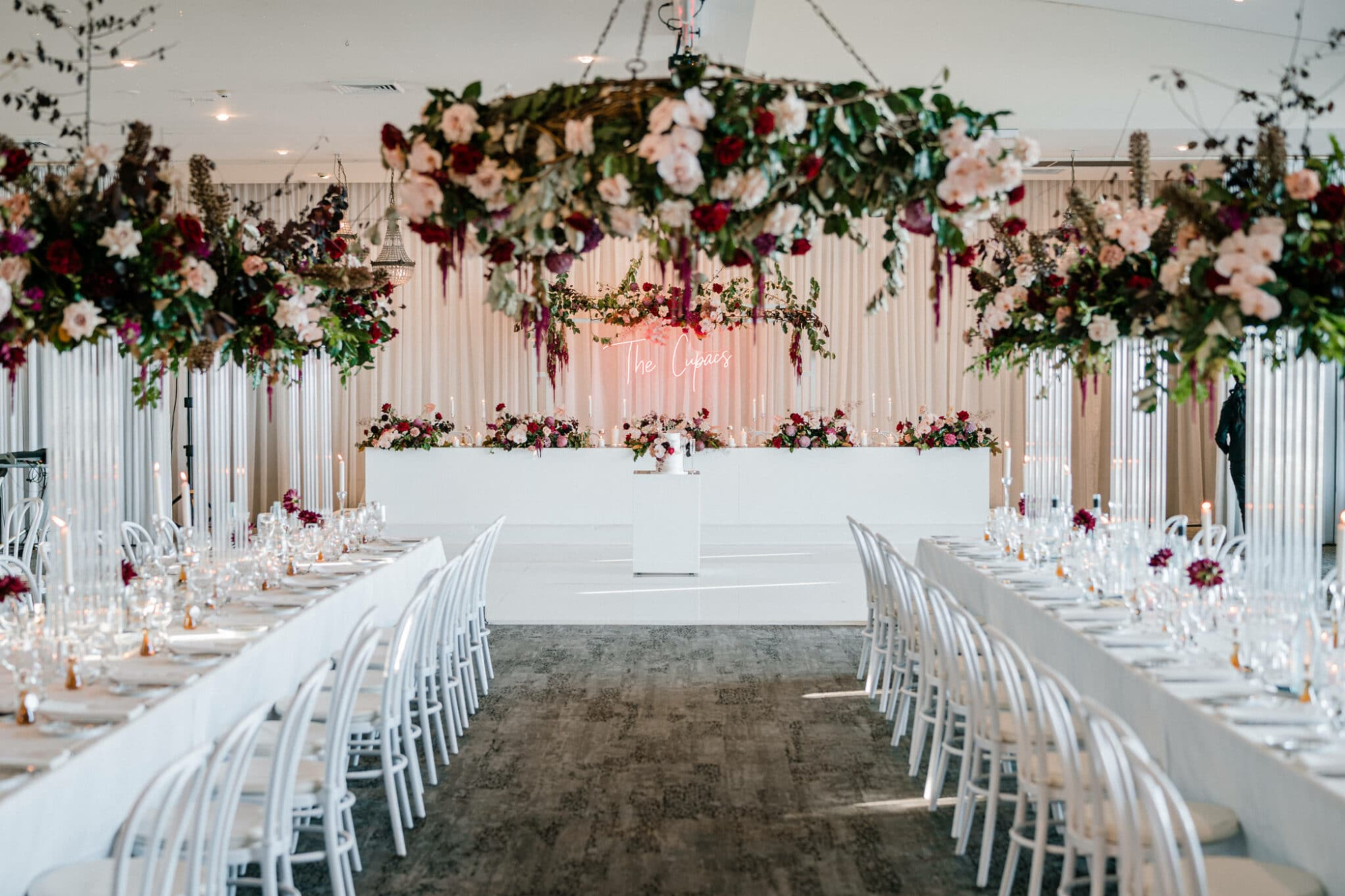 Elegant indoor wedding reception with long white tables, candles, and lush hanging floral arrangements.