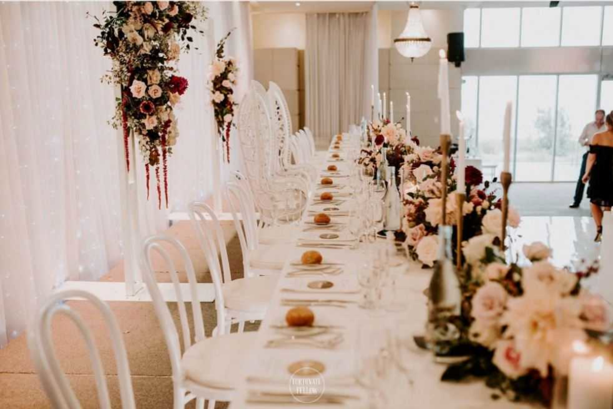 Elegant indoor wedding reception table with white chairs, tall candles, and lush floral arrangements in soft pink and burgundy tones.