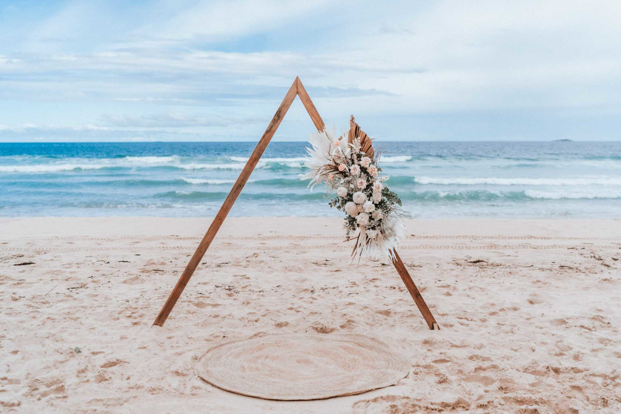 Wooden triangle wedding arch with boho florals set on a sandy beach by the ocean.