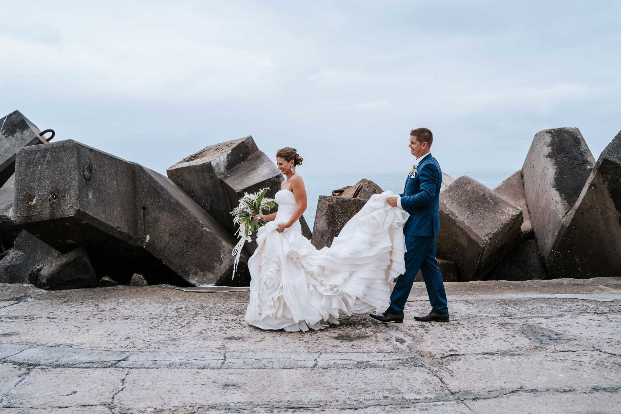 Bride and groom walk together by large concrete rocks near the sea, with the groom holding the bride’s flowing dress.
