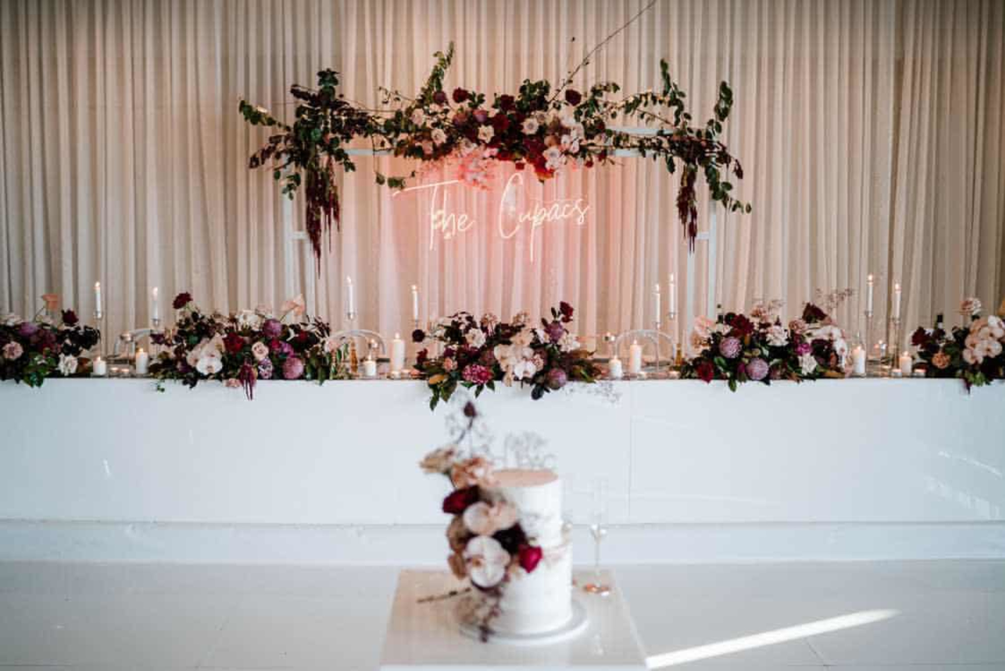 Romantic wedding head table with candles, abundant florals, neon sign backdrop, and a tiered cake in the foreground.