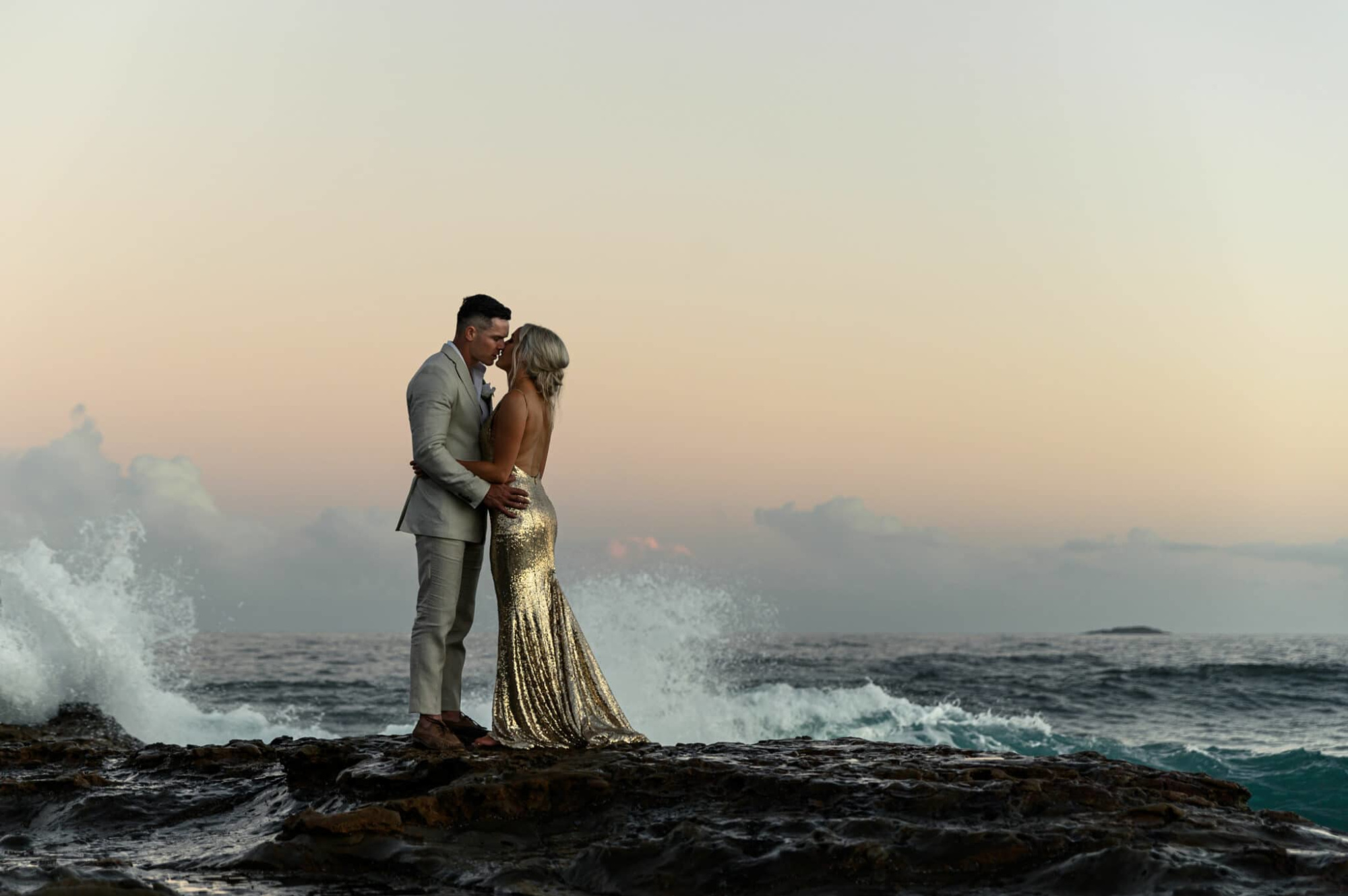 Romantic couple embracing on rocky shoreline at sunset with waves crashing behind them.