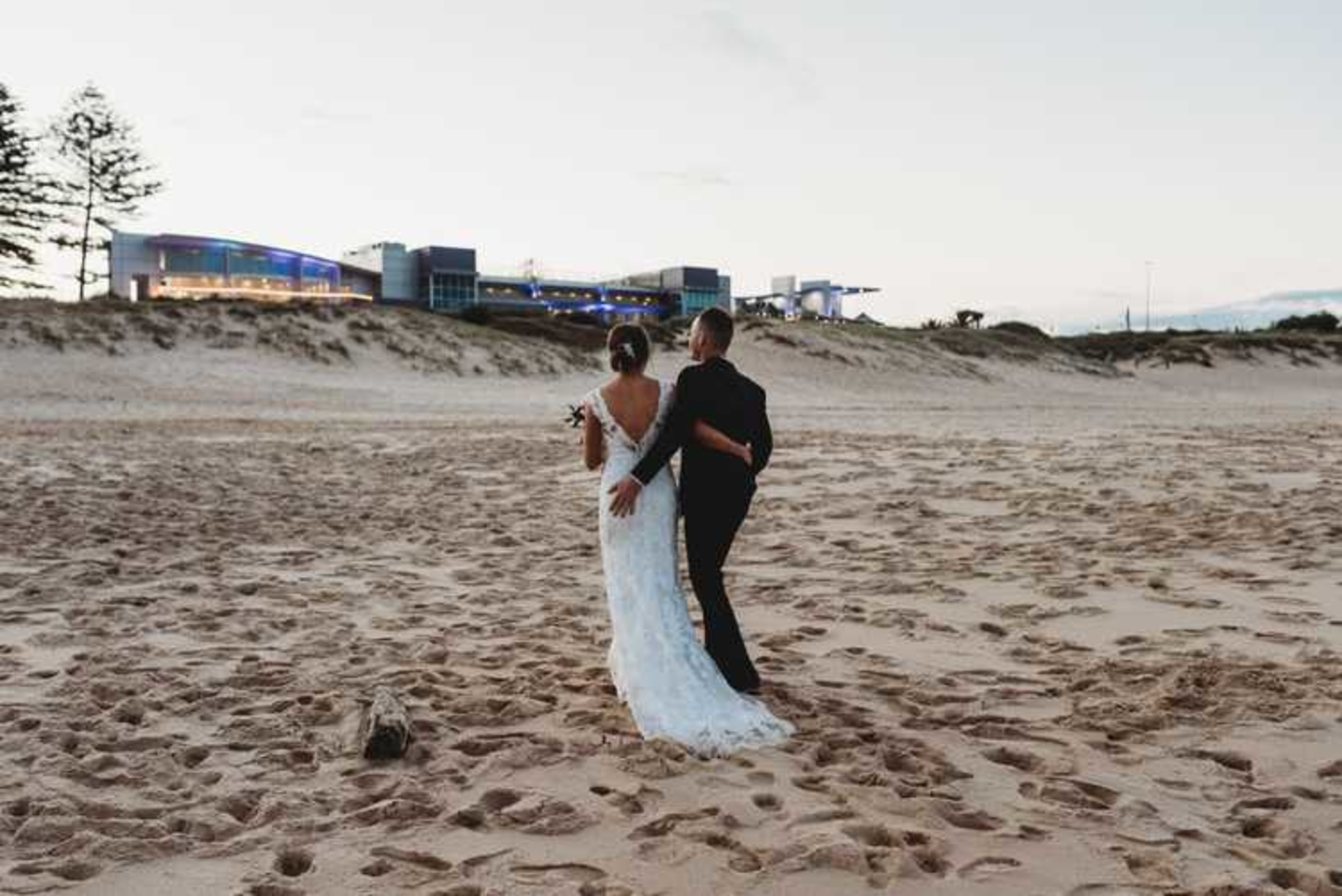 Bride and groom walk arm in arm along a sandy beach with a modern waterfront venue in the distance.