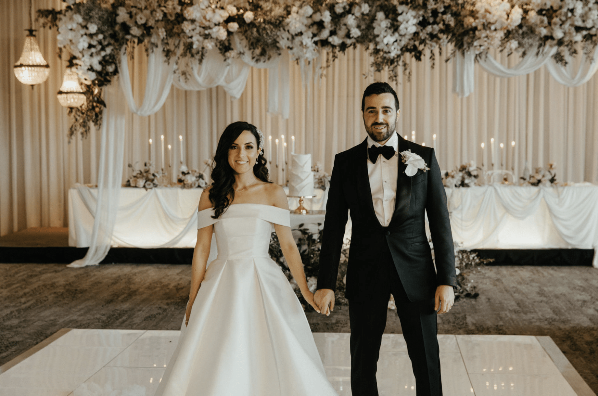Bride and groom holding hands in an elegant indoor reception with lush hanging florals and candlelit backdrop.