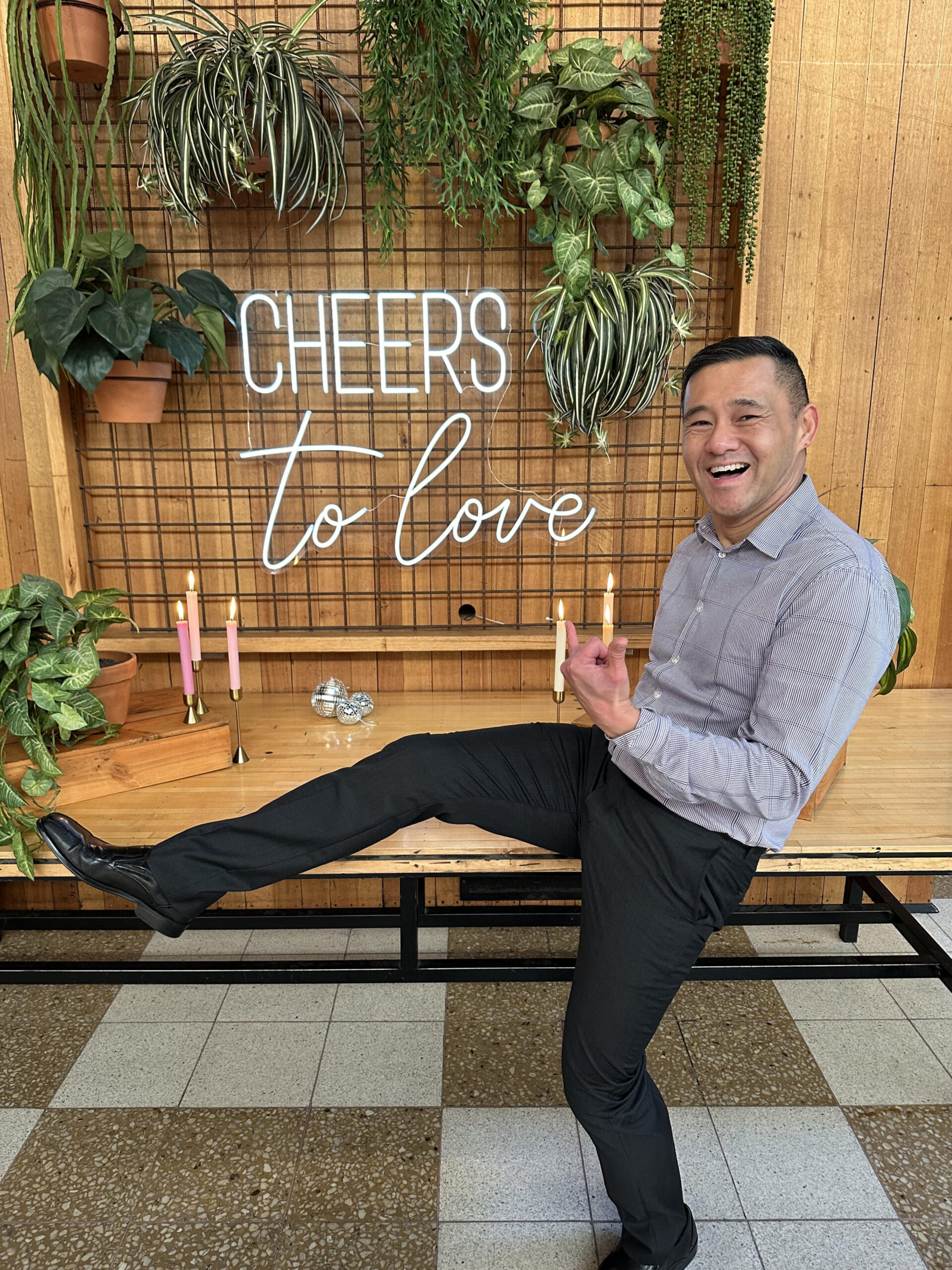 Smiling guest poses playfully in front of a neon 'Cheers to love' sign with greenery and candles.