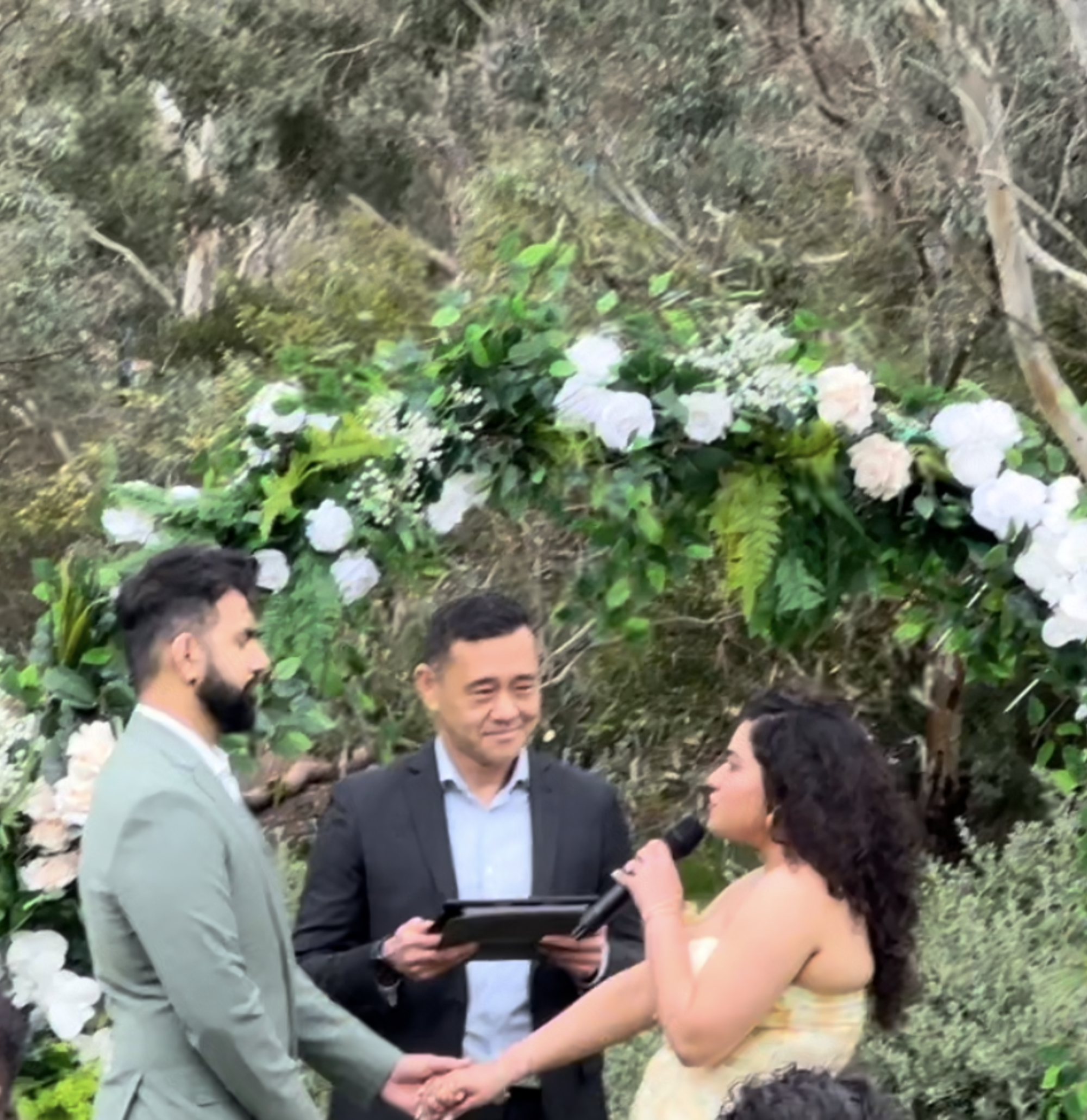Couple exchanging vows under a lush floral arch with an officiant at an outdoor garden wedding.