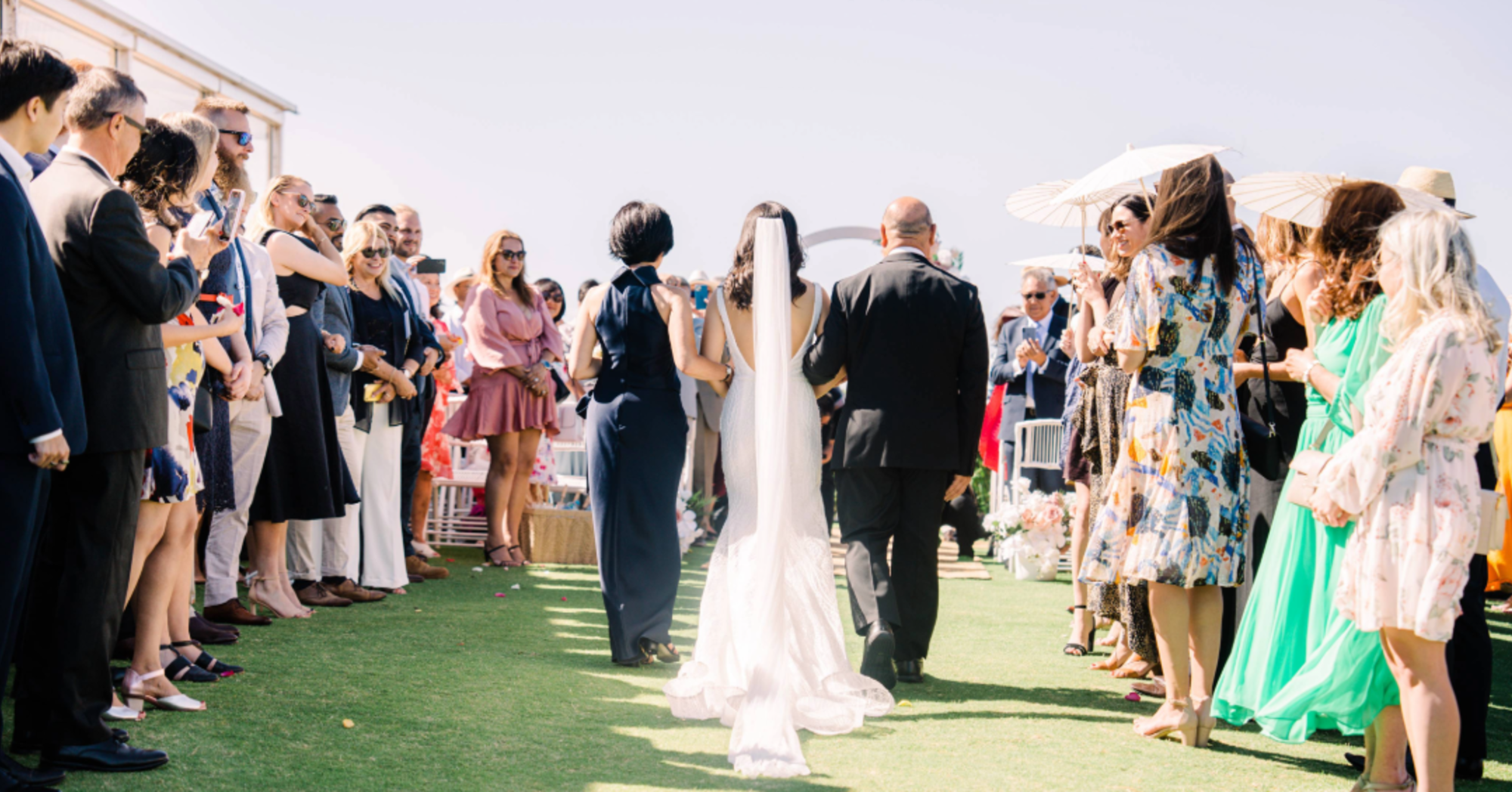 Bride walks down an outdoor aisle between rows of standing guests during a sunny wedding ceremony.