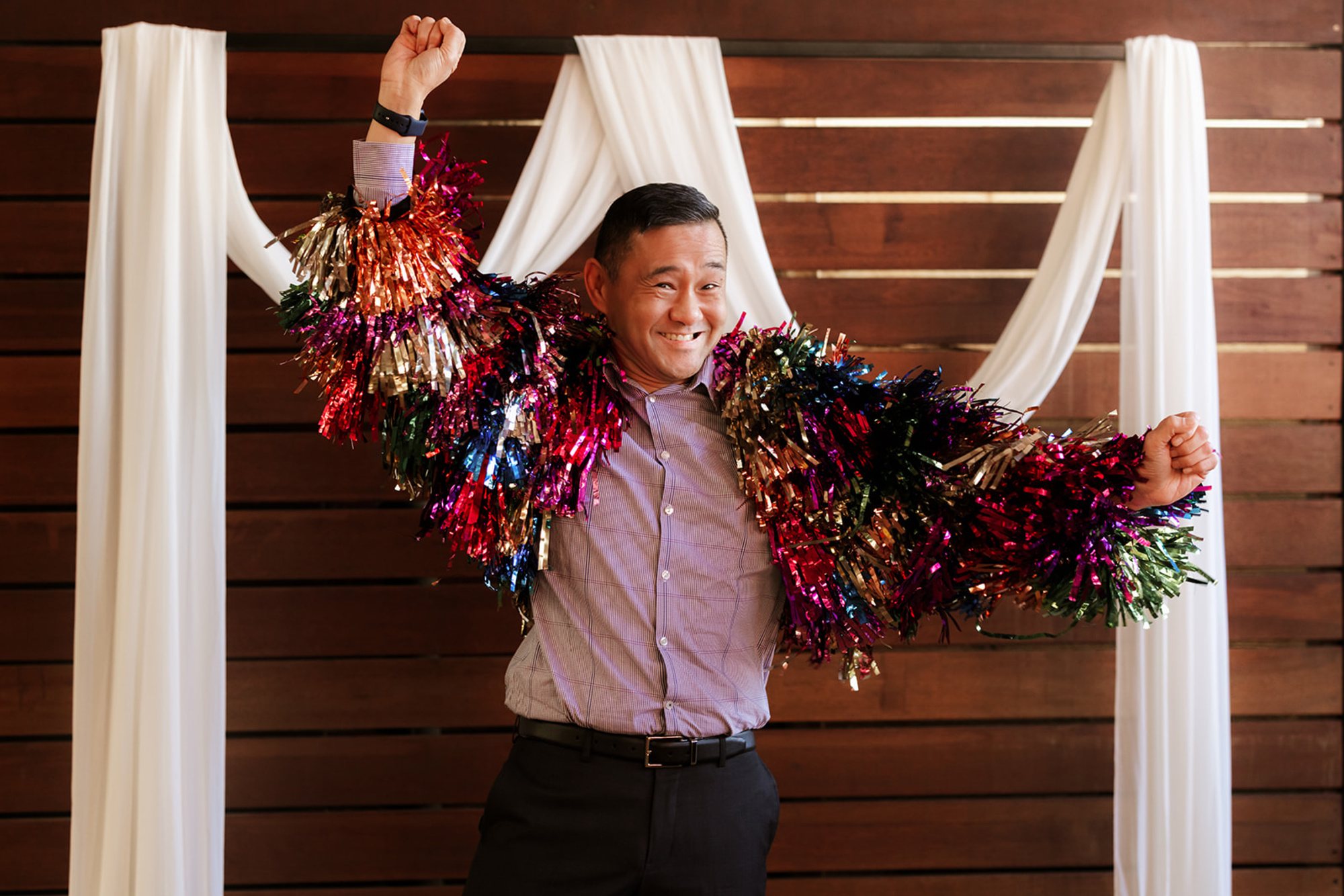 Smiling guest in a colorful tinsel jacket posing playfully in front of a draped wooden wedding backdrop.