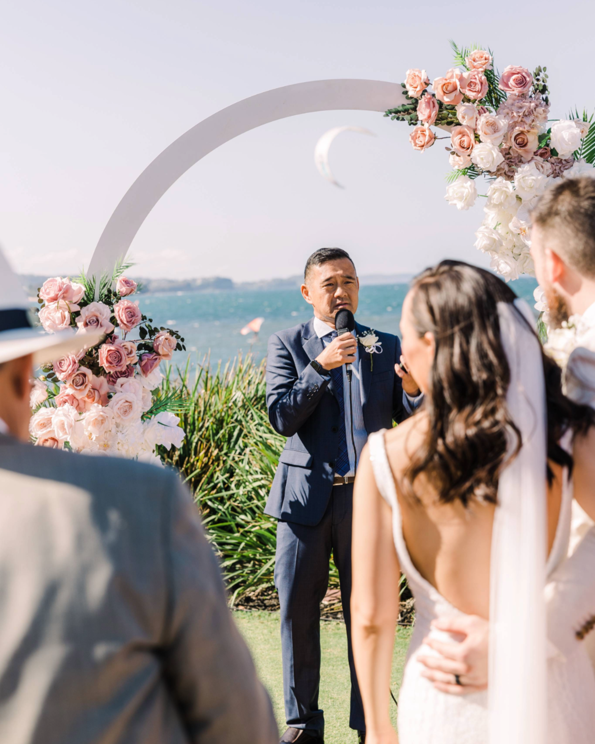 Officiant speaking at an outdoor waterfront wedding ceremony under a floral arch with the couple in the foreground.