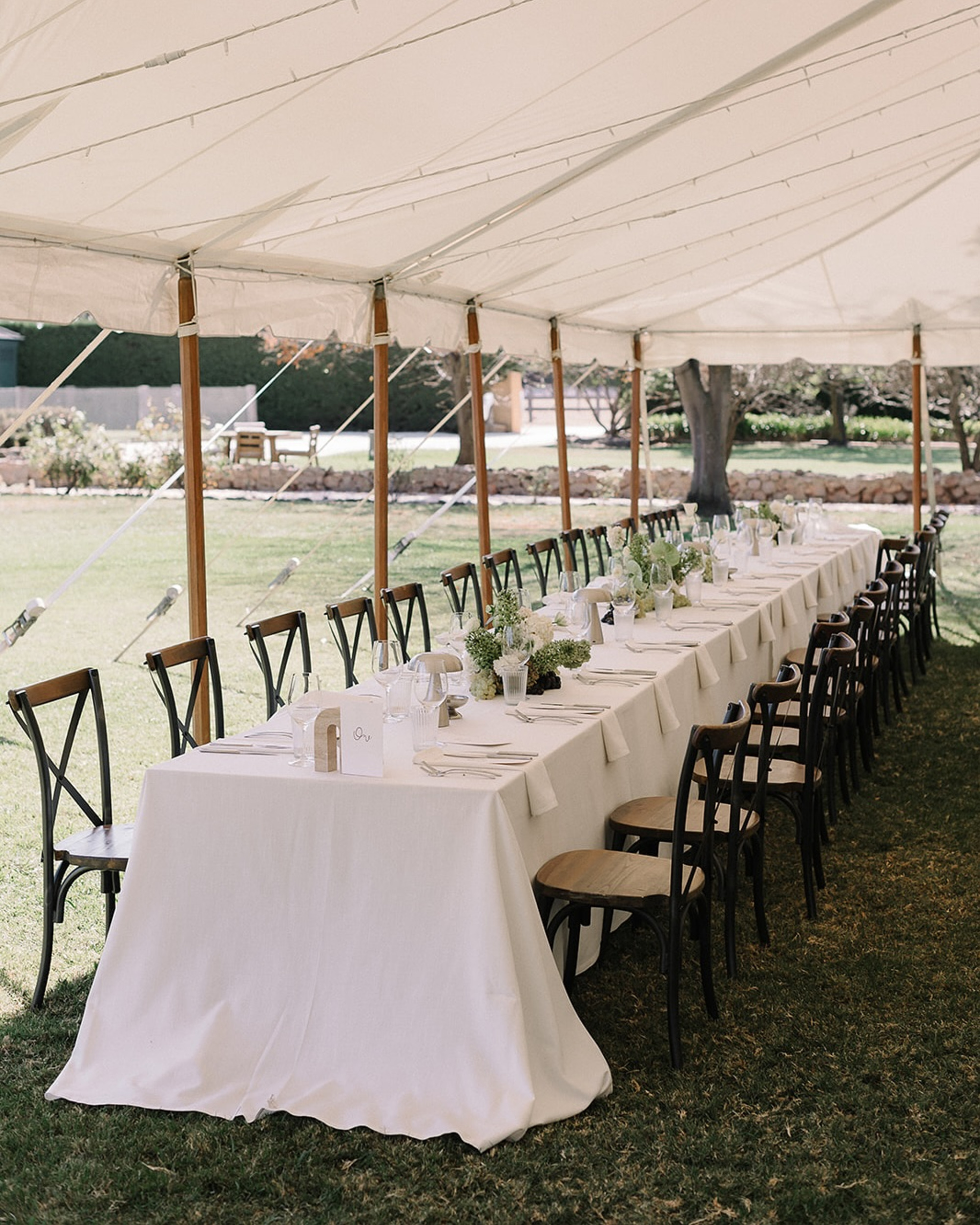 Long ivory-draped reception table set under a white tent on a grassy outdoor wedding lawn.