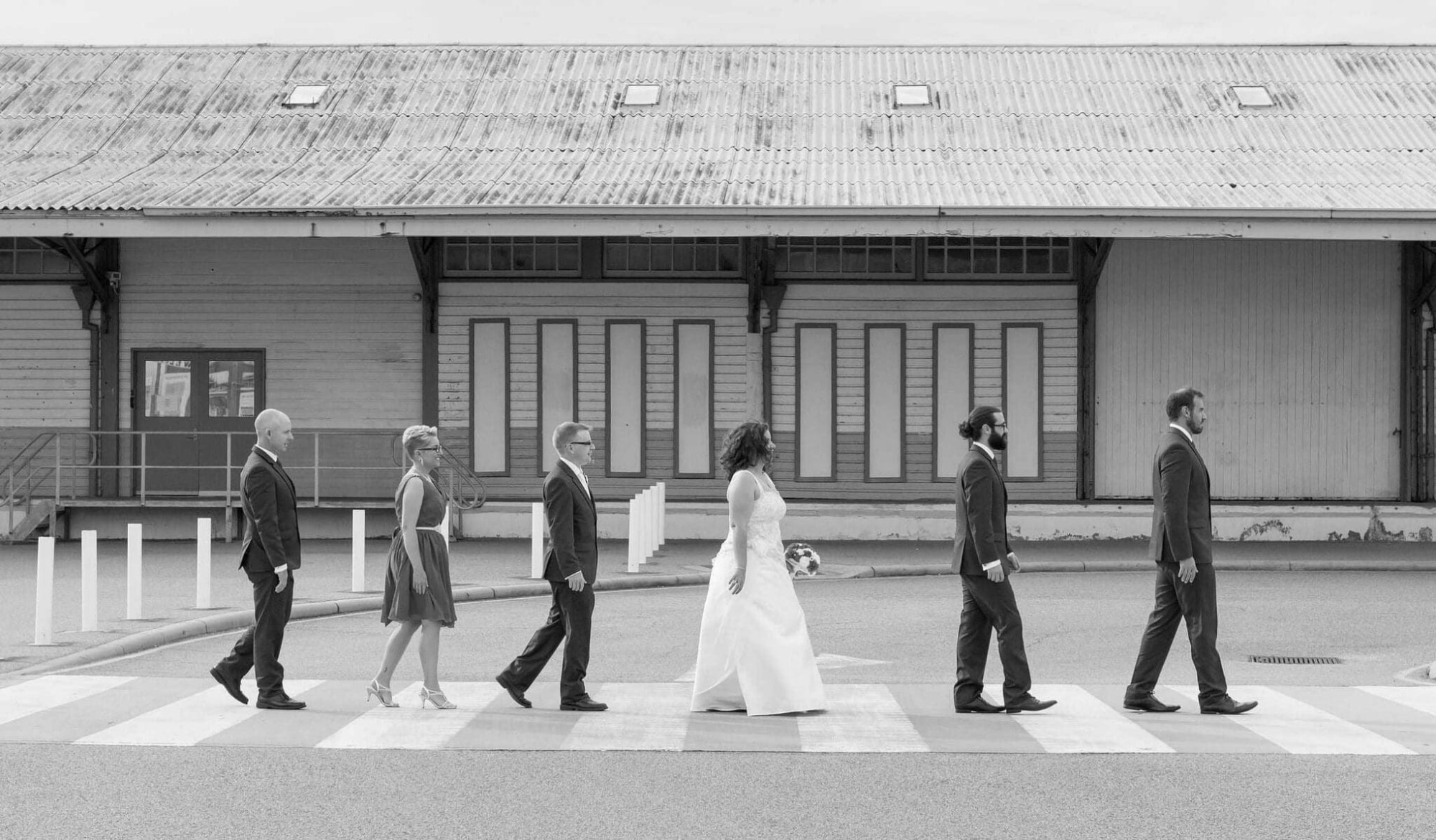 Wedding party walks in a line across a crosswalk in front of an industrial building in a black and white photo.