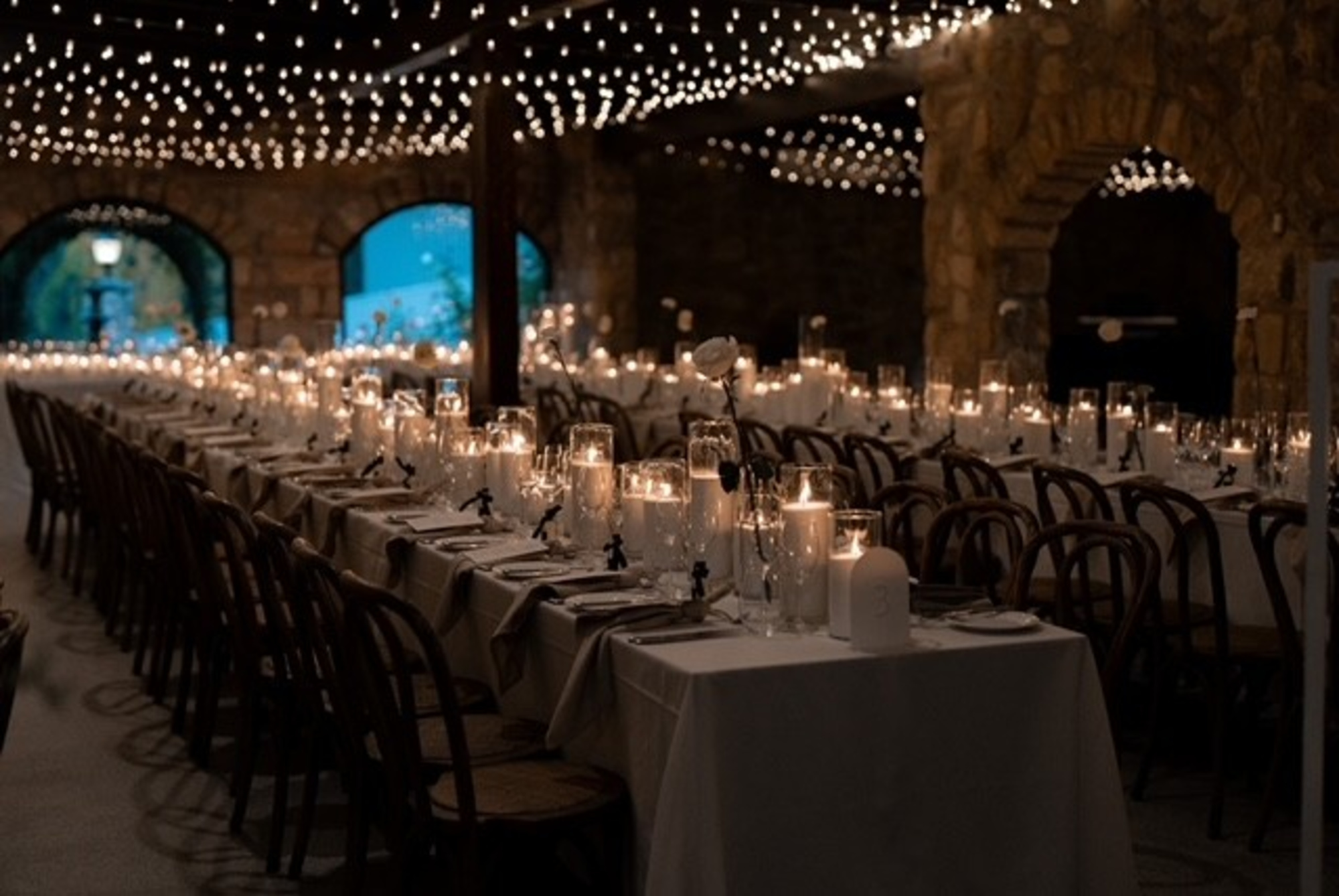 Long candlelit wedding reception tables under twinkling string lights in a stone arch venue at dusk.