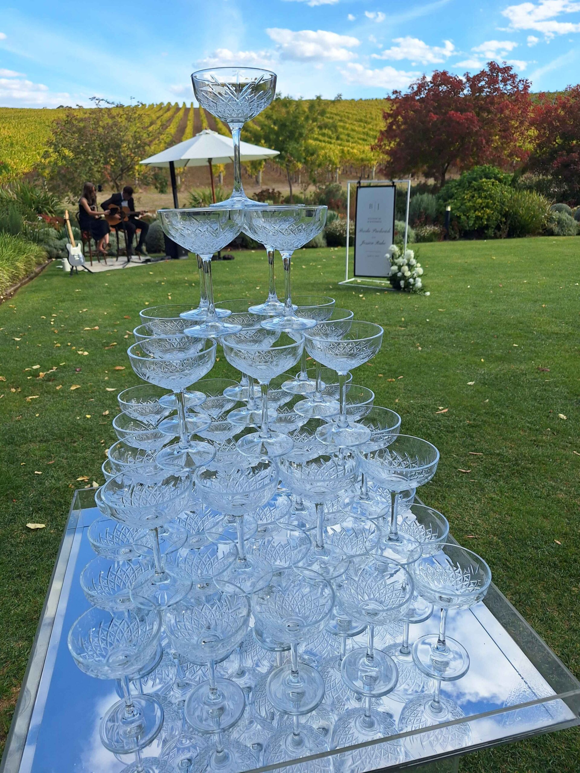 Crystal champagne tower set on a mirrored table at an outdoor vineyard wedding reception.