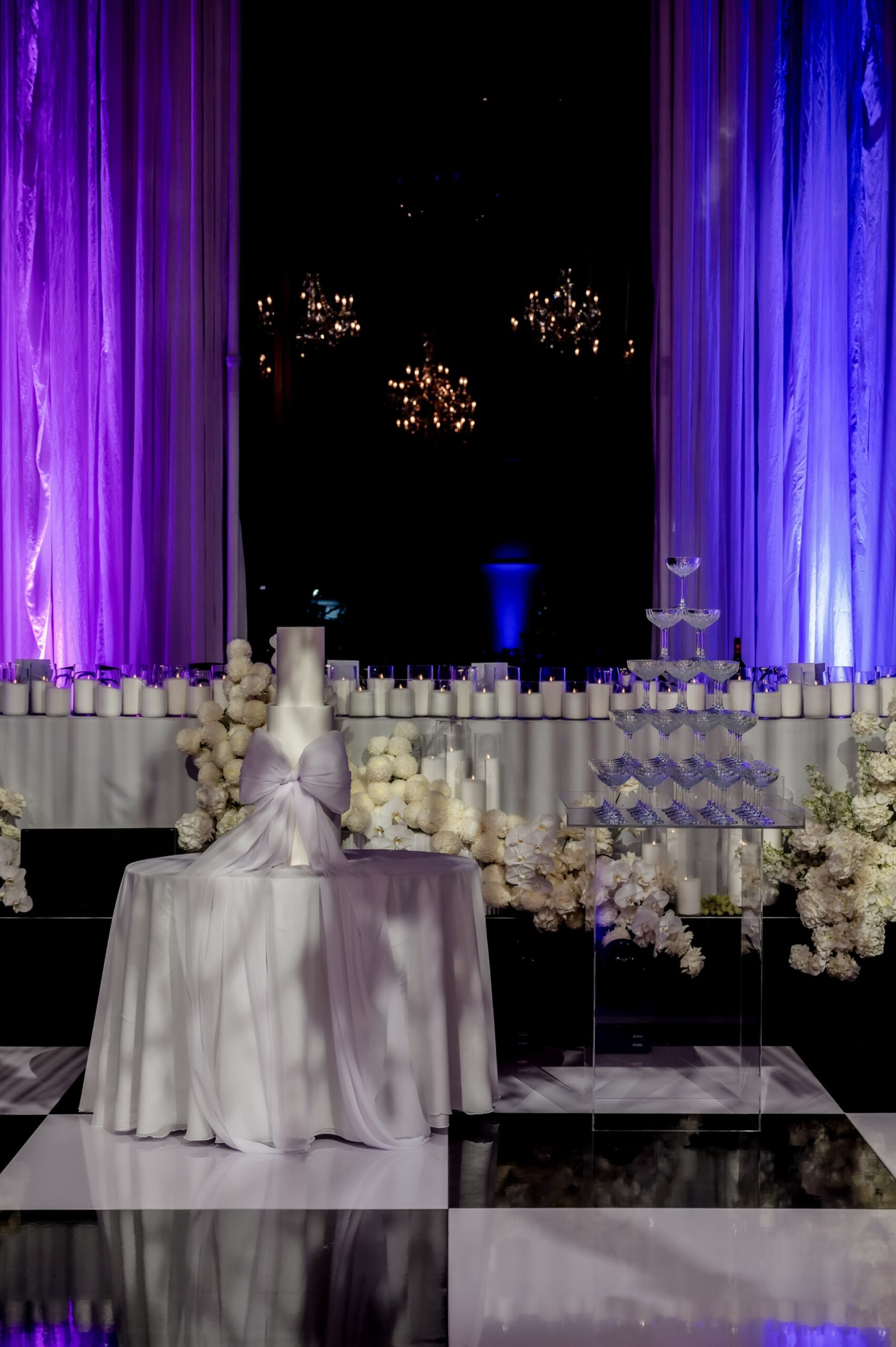 Elegant wedding cake and champagne tower surrounded by white flowers and candles with purple uplighting.