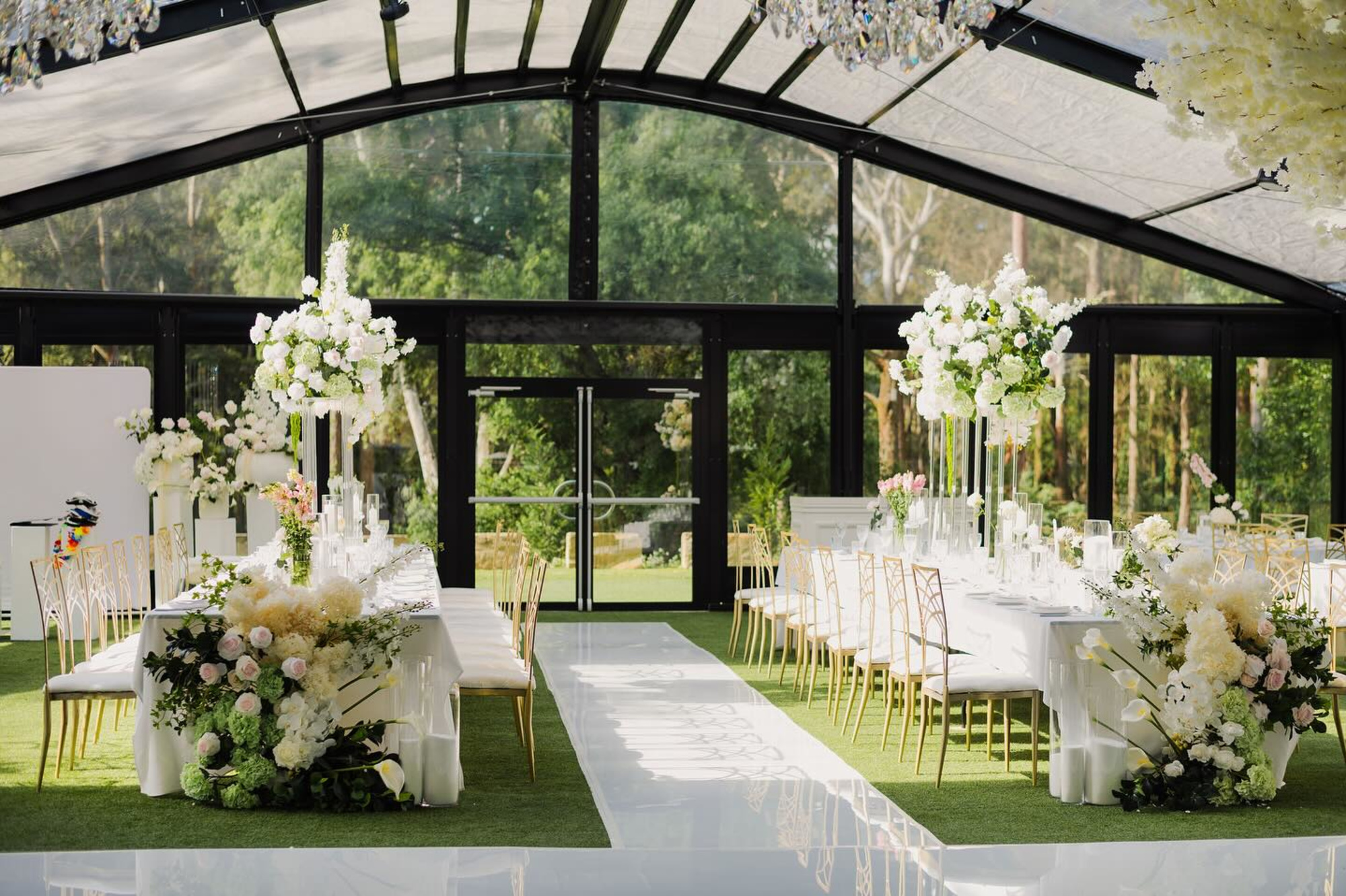 Elegant glass marquee wedding reception with long white tables, tall floral centerpieces, and gold chairs overlooking a lush garden.