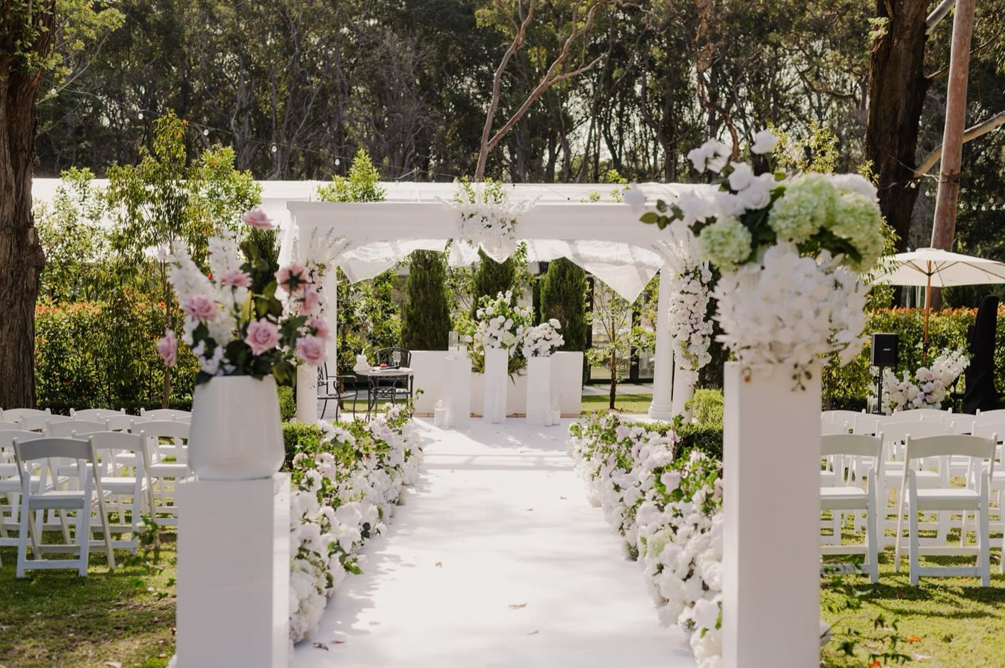 Outdoor garden wedding ceremony setup with white floral aisle and elegant archway.