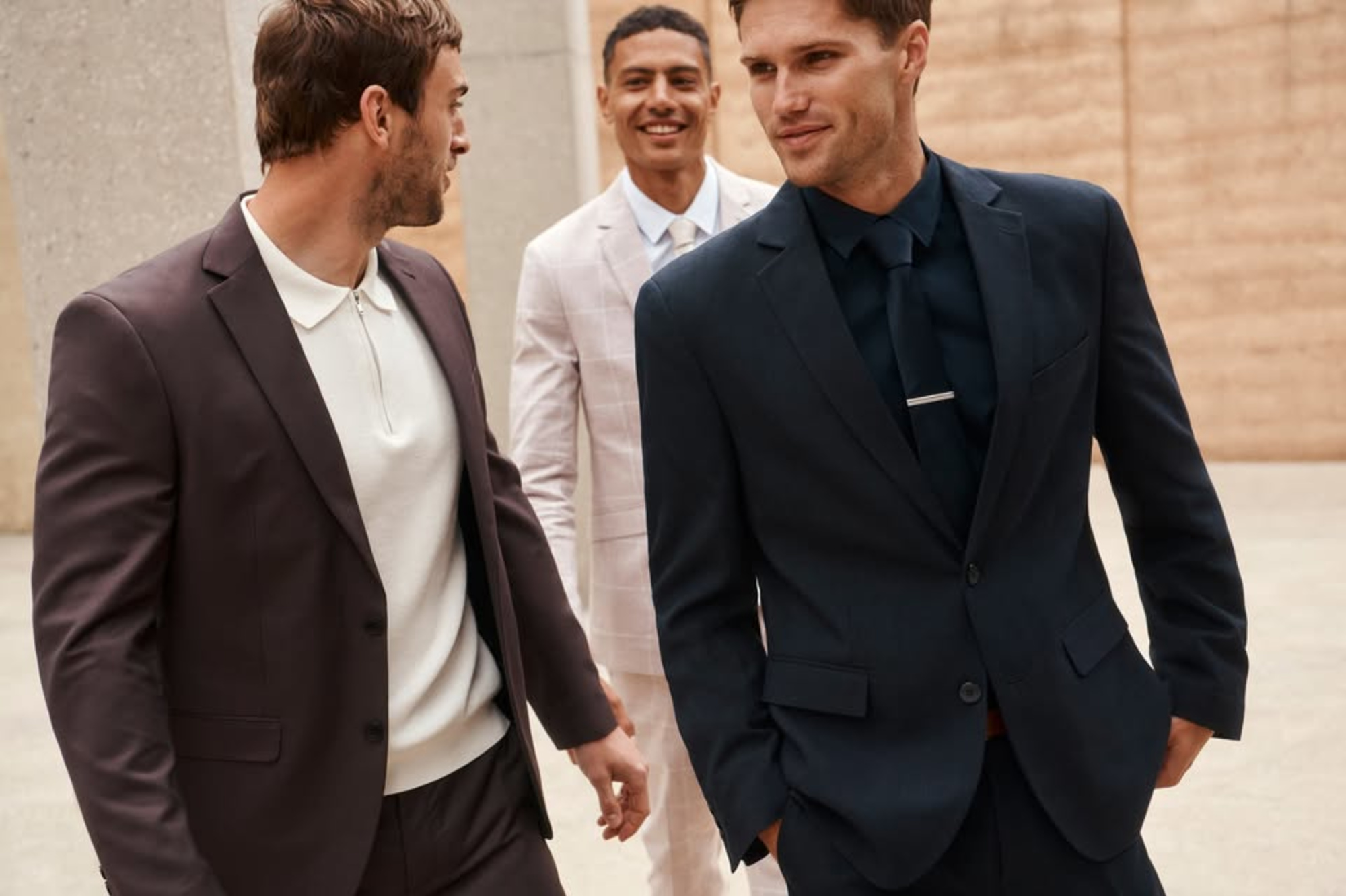 Three men in stylish suits walk together outdoors, showcasing modern wedding groomsmen attire.