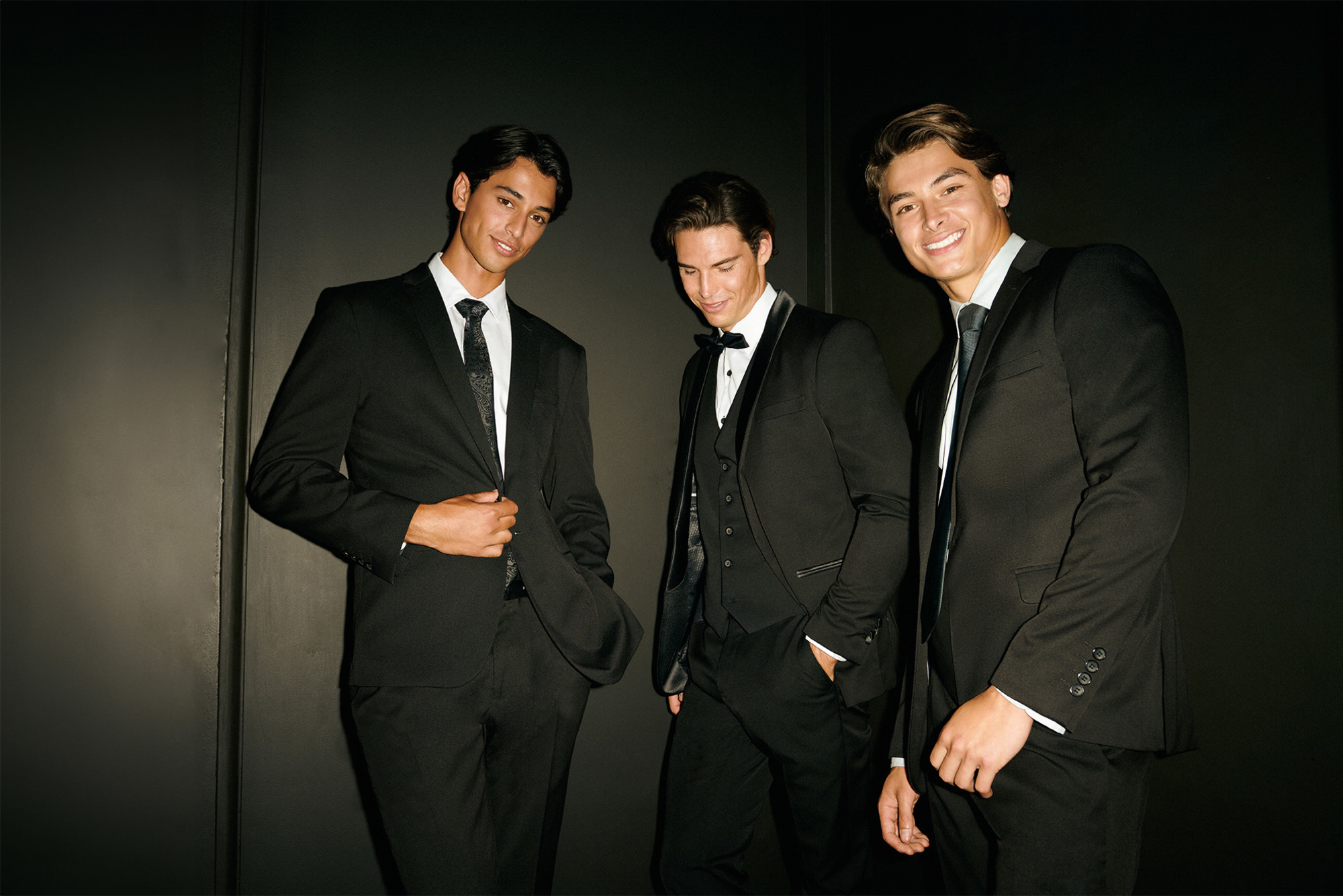 Three young men in sleek black suits and tuxedos pose against a dark backdrop for modern wedding attire.