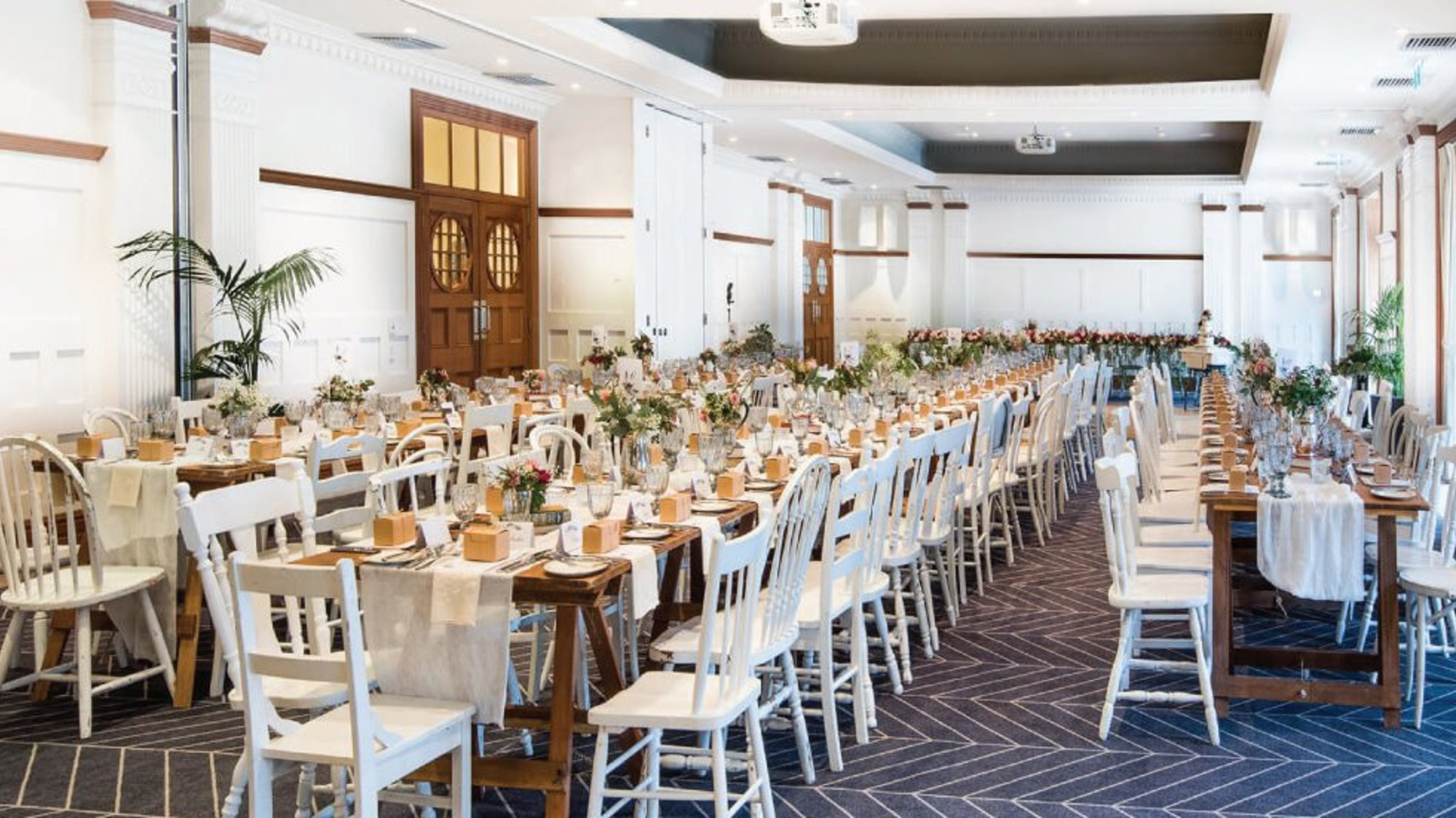 Bright indoor wedding reception hall with long wooden tables, white chairs, and elegant floral centerpieces.