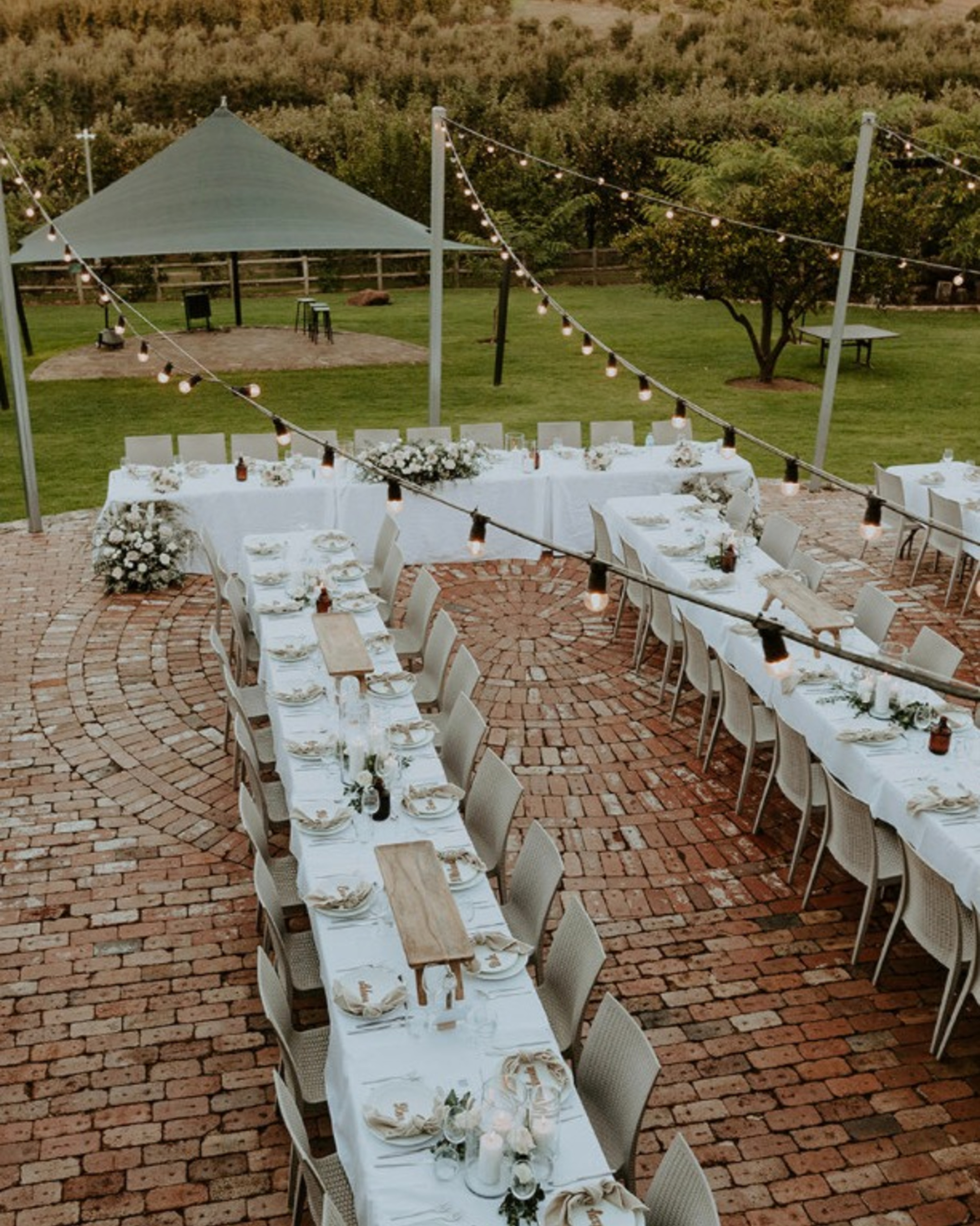 Outdoor wedding reception setup with long white-draped tables on a brick patio under string lights in a green garden.