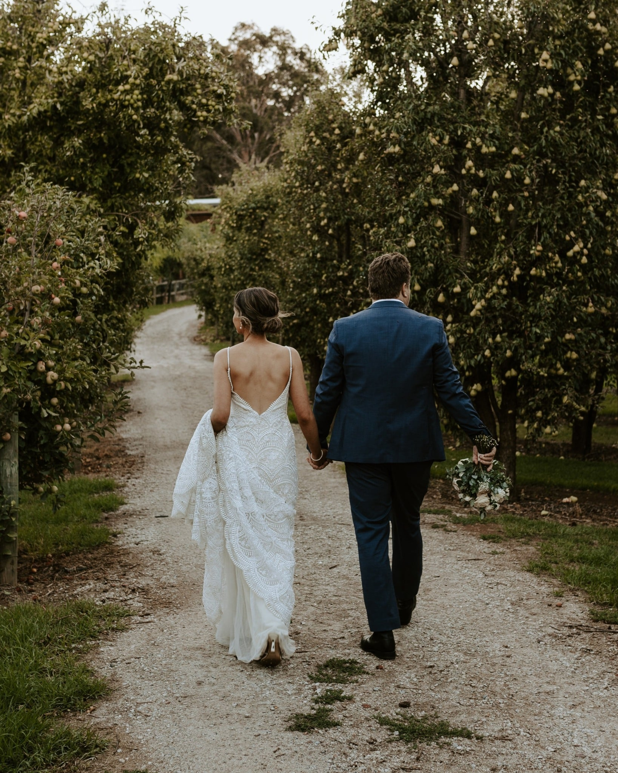 Newlywed couple walks hand in hand down a rustic orchard path after their outdoor wedding ceremony.