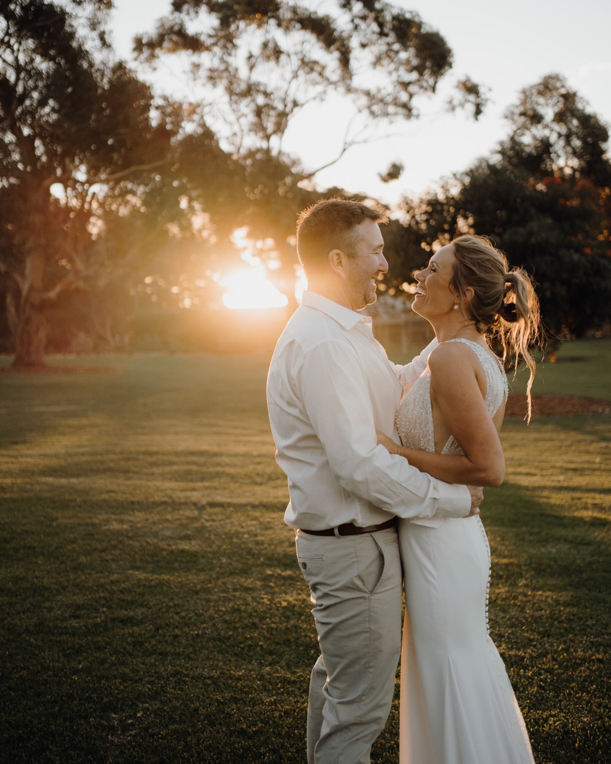 A bride and groom embrace and smile at each other during sunset in a grassy outdoor setting.