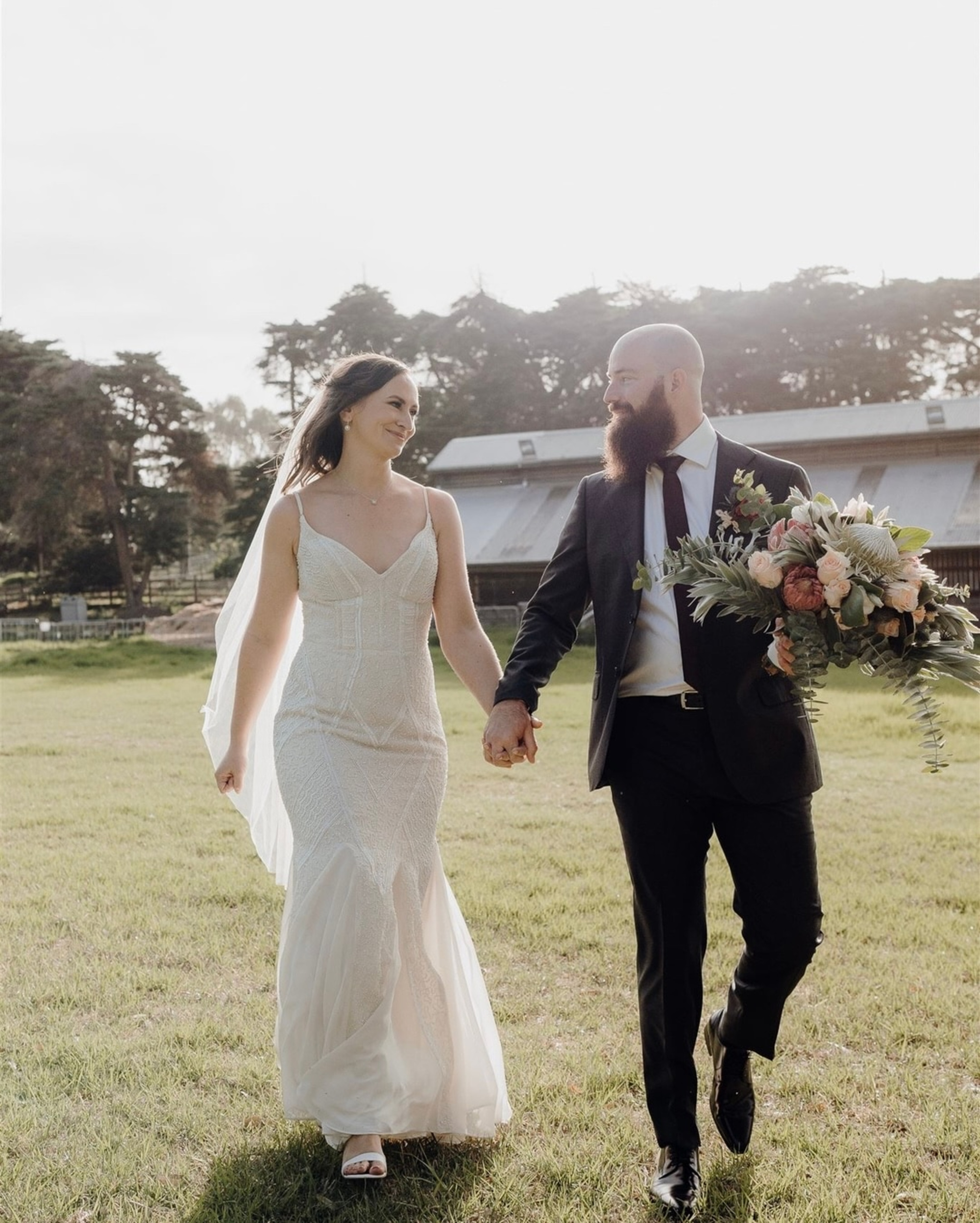 Newlywed couple holding hands and walking through a sunlit grassy field with a rustic barn in the background.