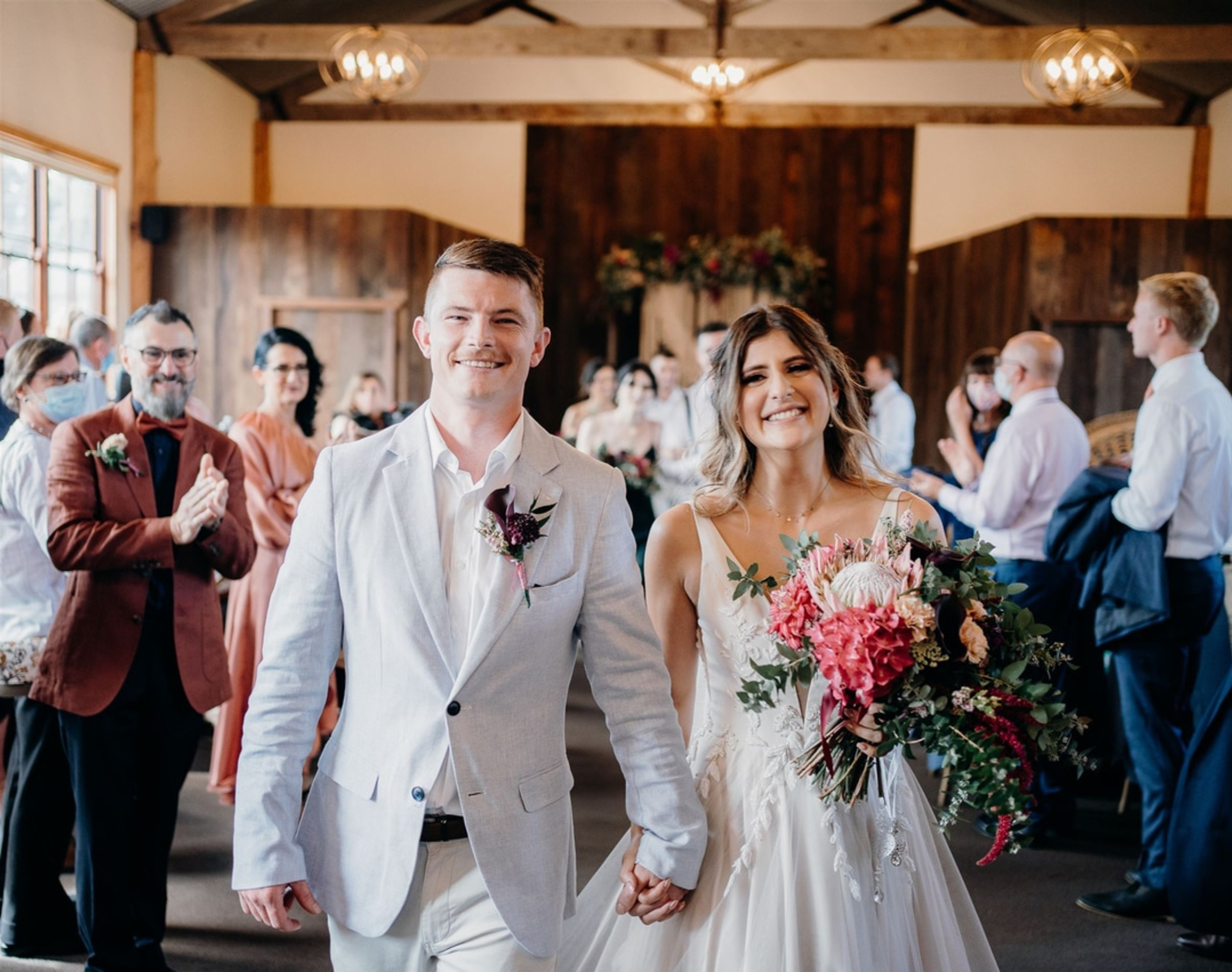 Smiling bride and groom walk hand in hand down the aisle of a rustic indoor wedding surrounded by applauding guests.