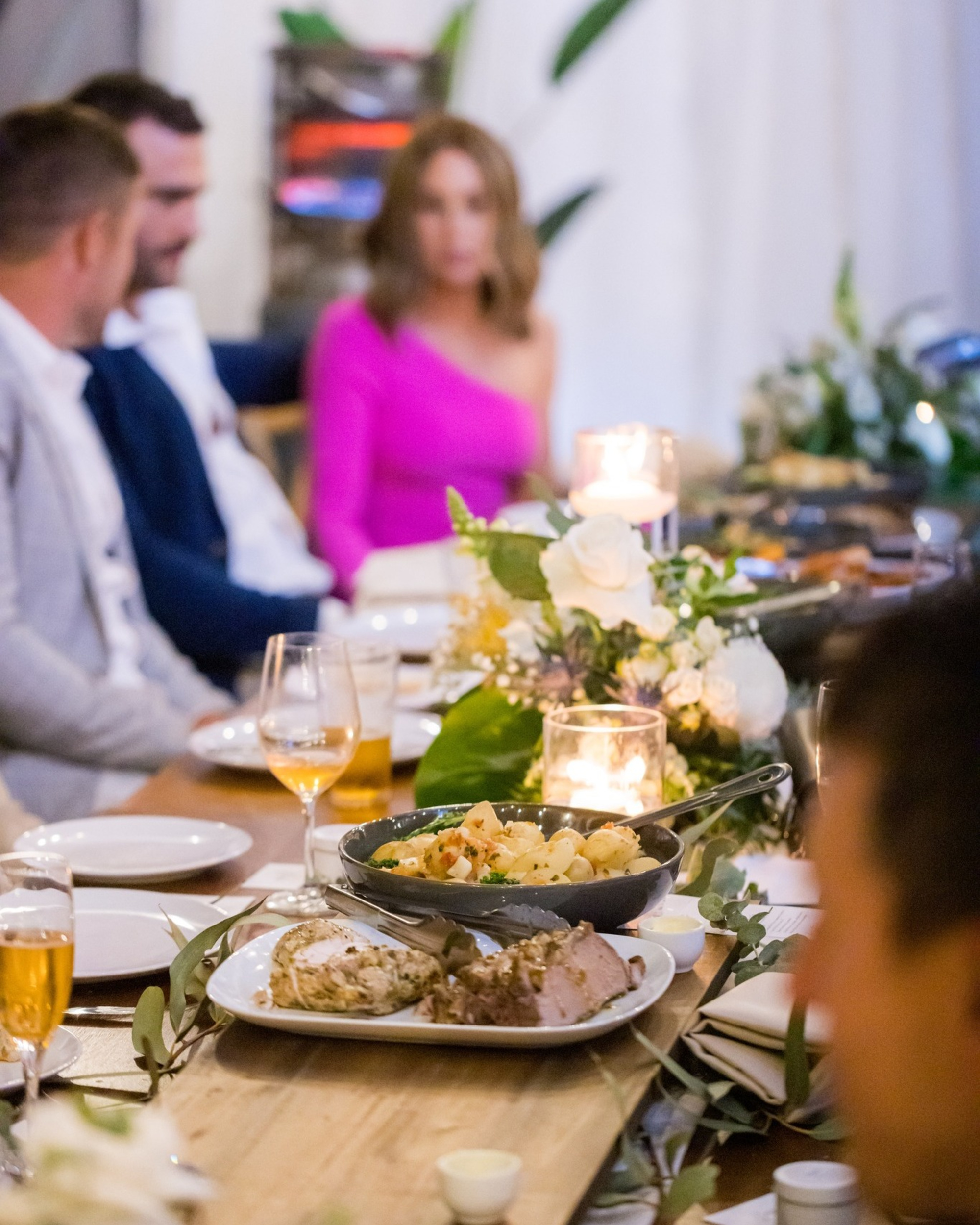 Candlelit wedding reception table with plated food, floral centerpieces, and guests seated and talking.