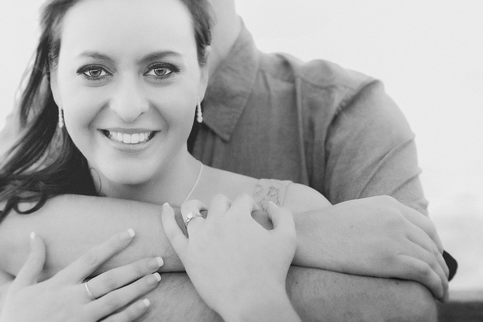 Close-up black and white portrait of a smiling woman embraced by her partner, showcasing her engagement ring.