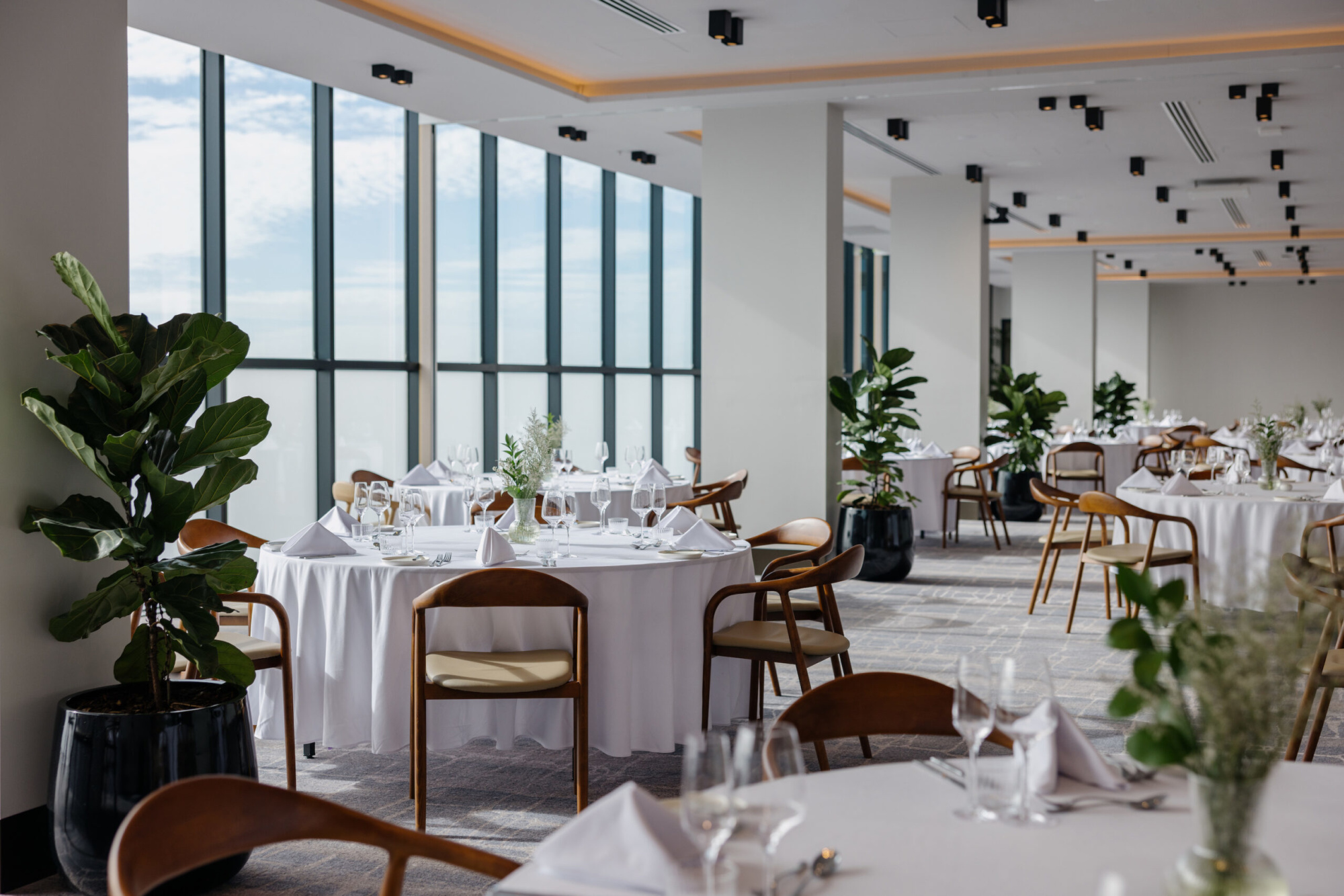 Modern light-filled wedding reception venue with round tables, white linens, and wooden chairs by large windows.