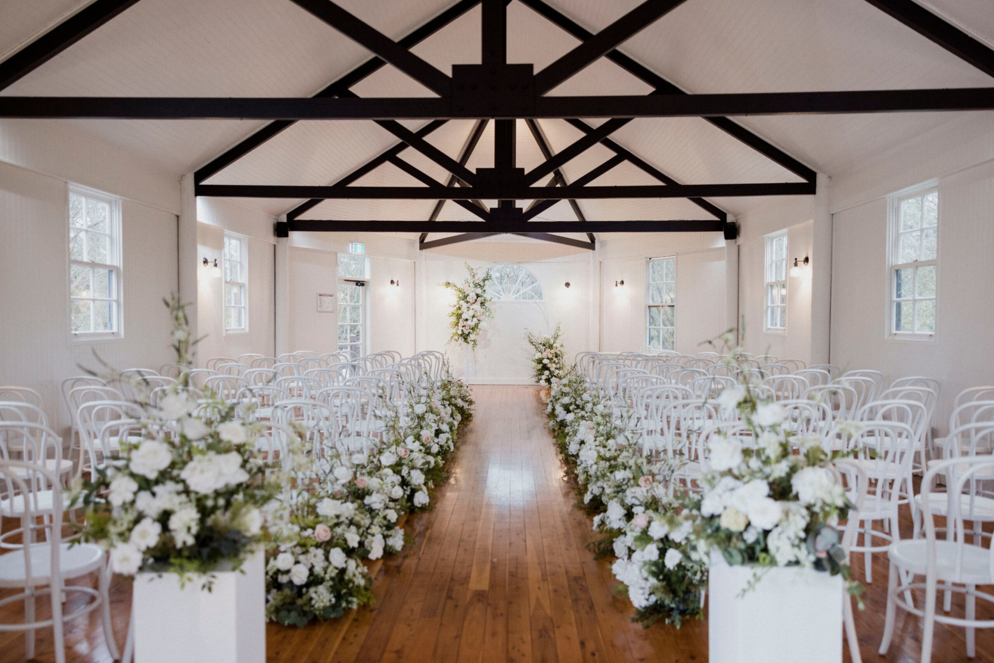 Bright indoor wedding ceremony setup with white chairs and lush floral arrangements lining a wooden aisle.