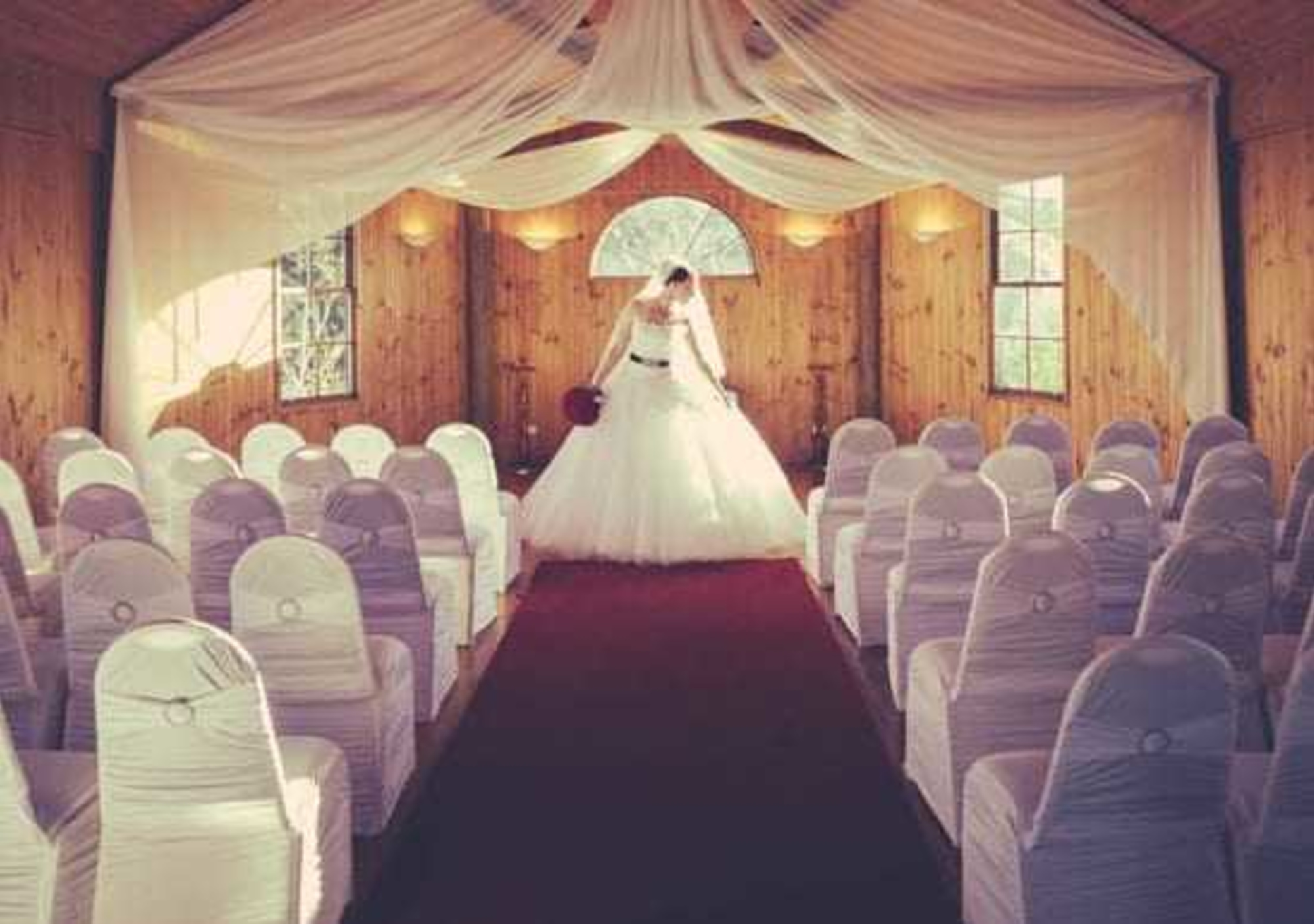 Bride stands at the front of a draped wooden chapel with white covered chairs lining a red aisle.