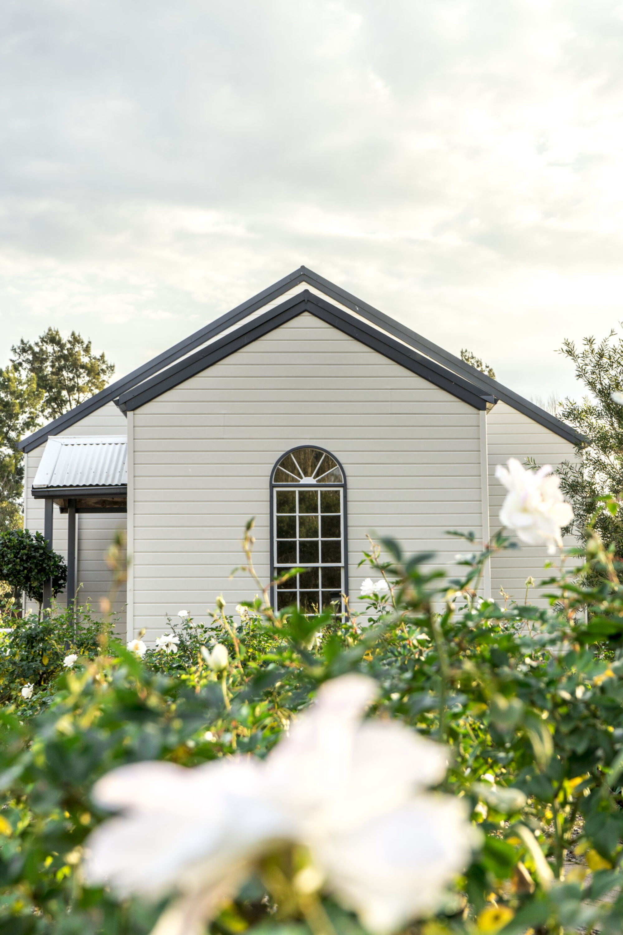 Small light-colored chapel surrounded by blooming garden flowers under a soft cloudy sky.