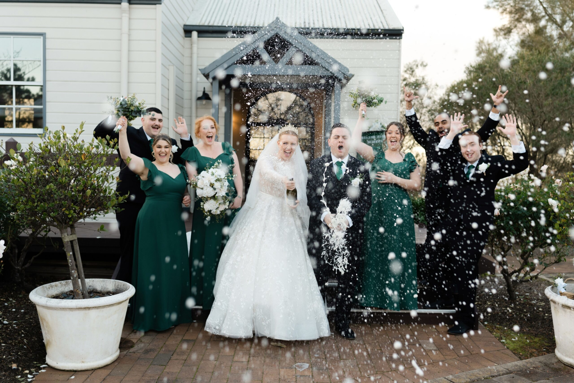 Joyful wedding party pops champagne outside a chapel, dressed in green gowns and black suits.